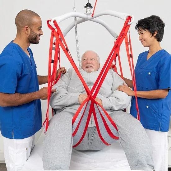 Two healthcare workers assisting a person in a ceiling lift sling. Red straps, blue scrubs, indoor setting.