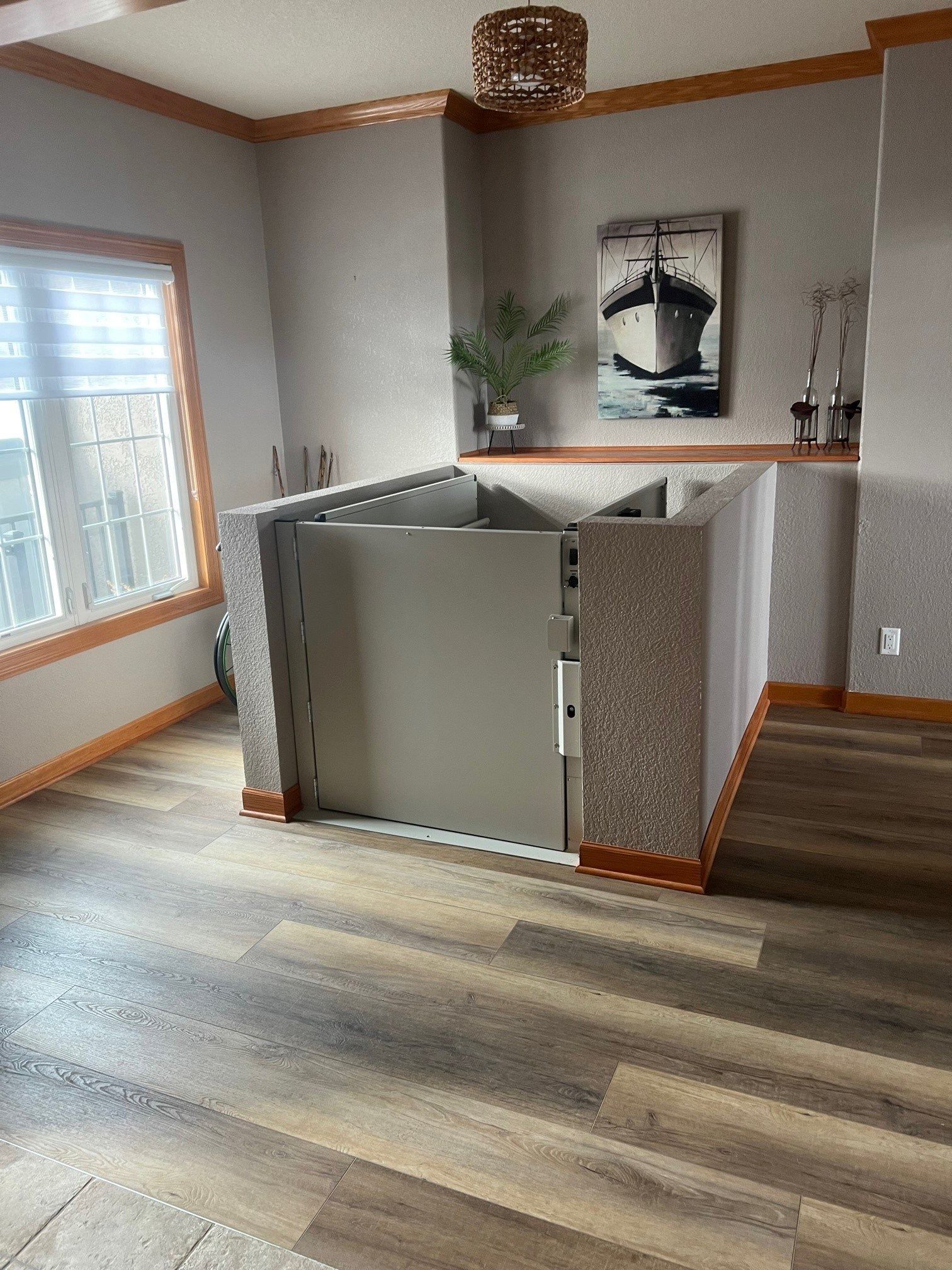 Indoor residential vertical platform lift, light grey, with textured columns. Wooden floor, window, artwork.