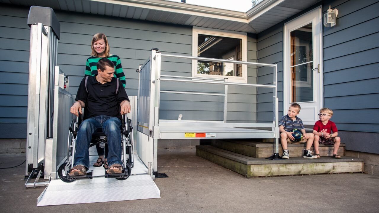 Person in wheelchair using a vertical platform lift with a family; house exterior.