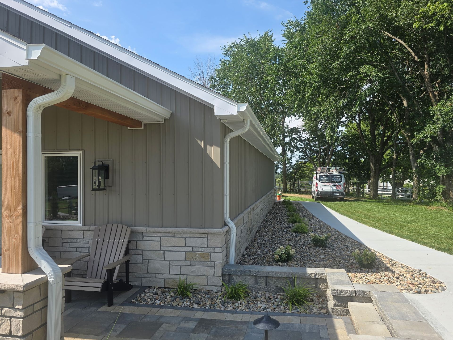 House exterior with stone foundation, front porch chairs, and a sloped rock-lined walkway beside trees.