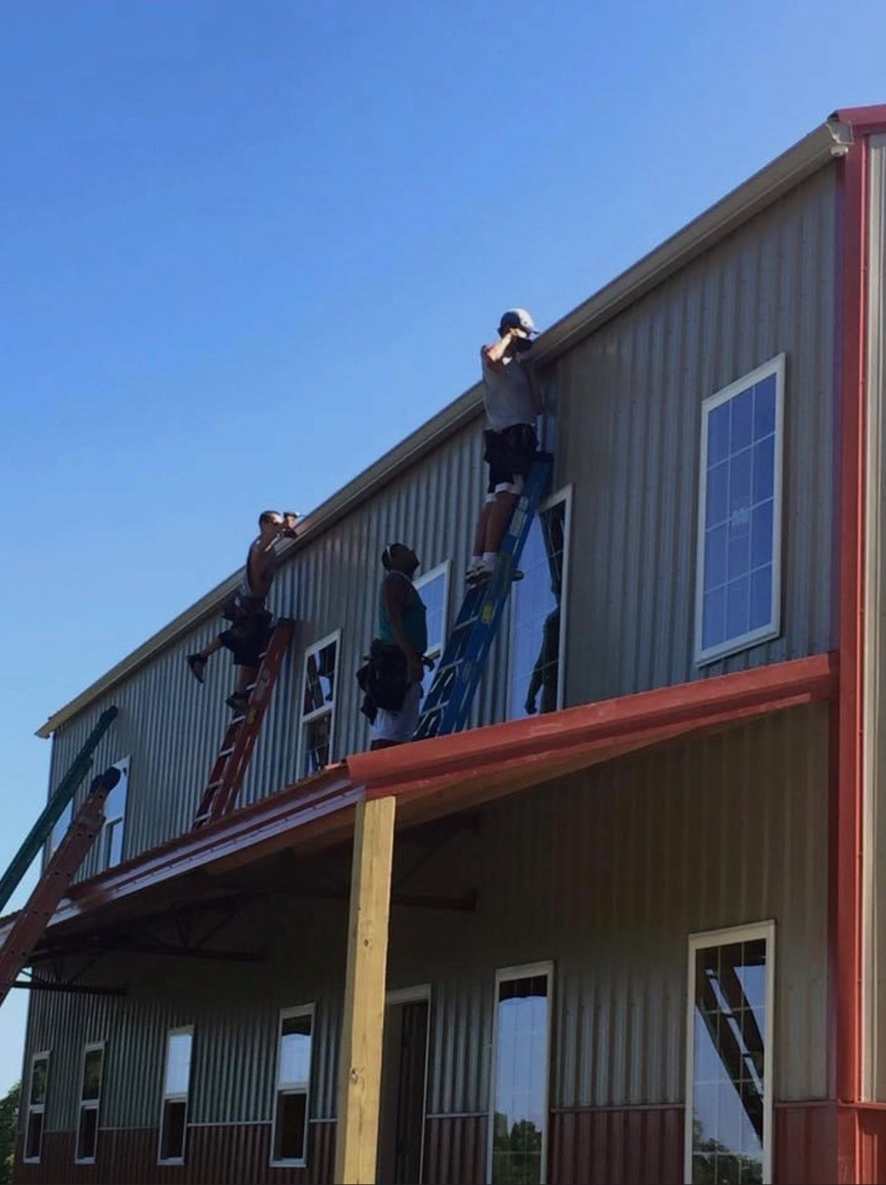 Three workers on ladders installing siding on a metal building with a red awning. Sunny day, blue sky.
