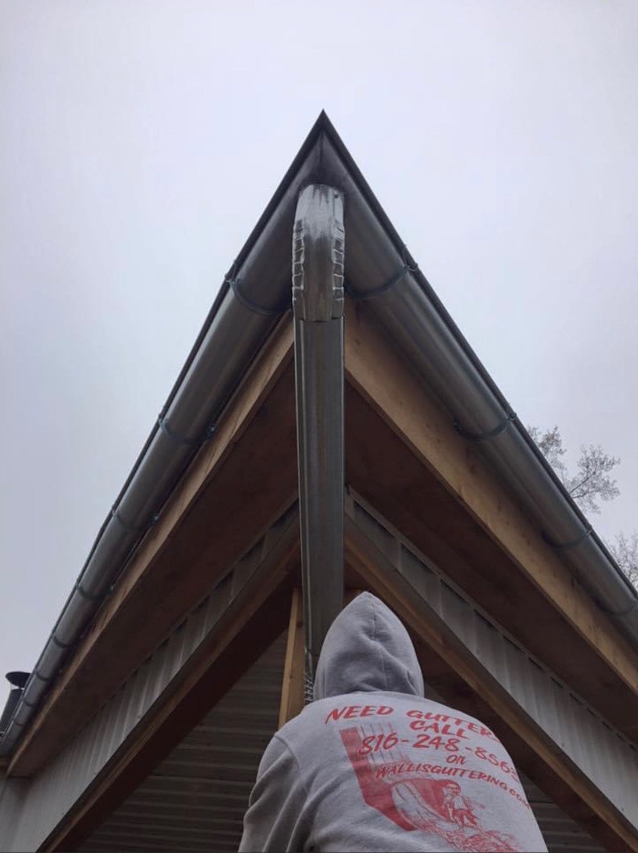 Person in a gray hoodie standing under a triangular roof with gutters. Overcast sky.