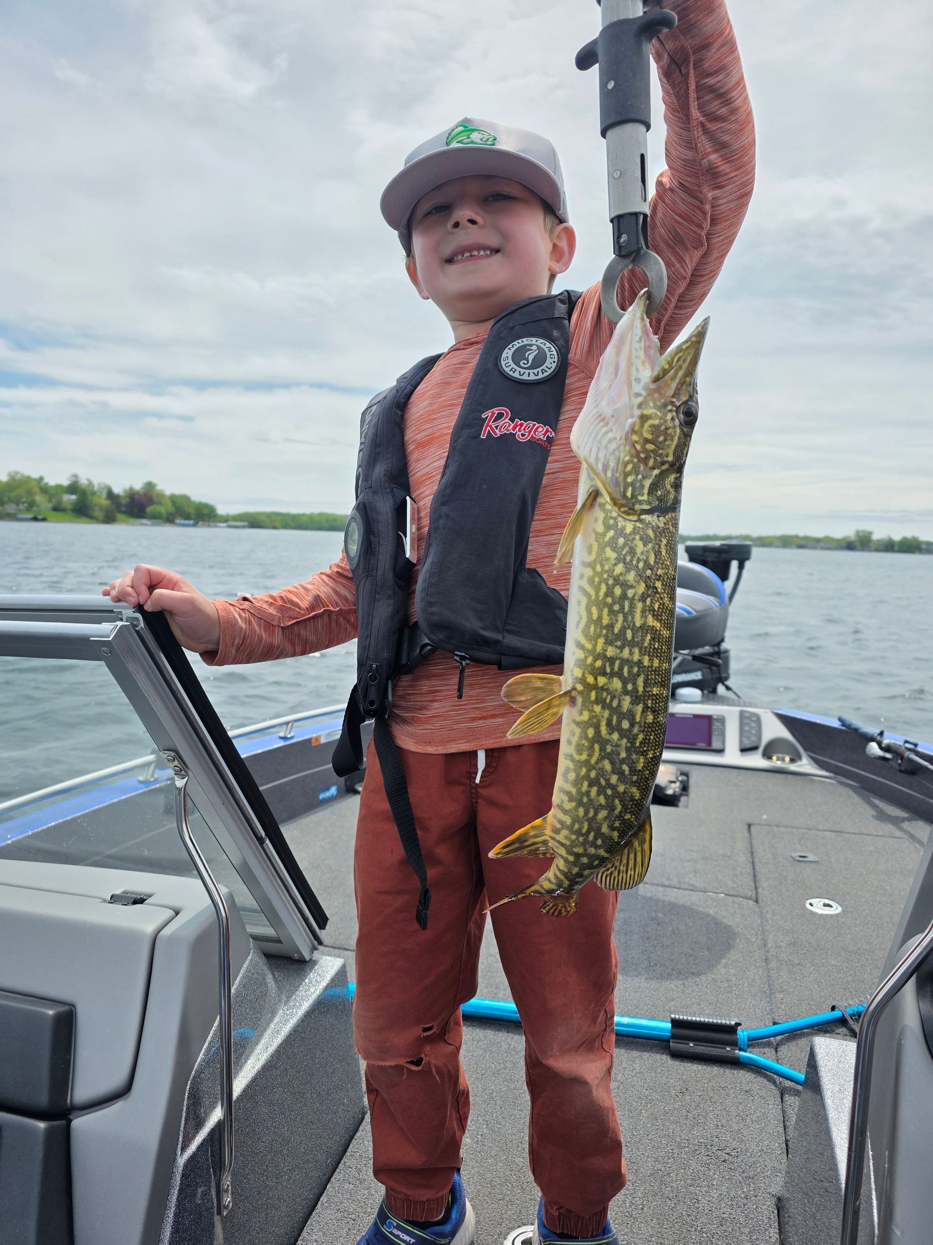 Child on a boat holding up a large fish, wearing brown rain gear and a cap on a cloudy day.