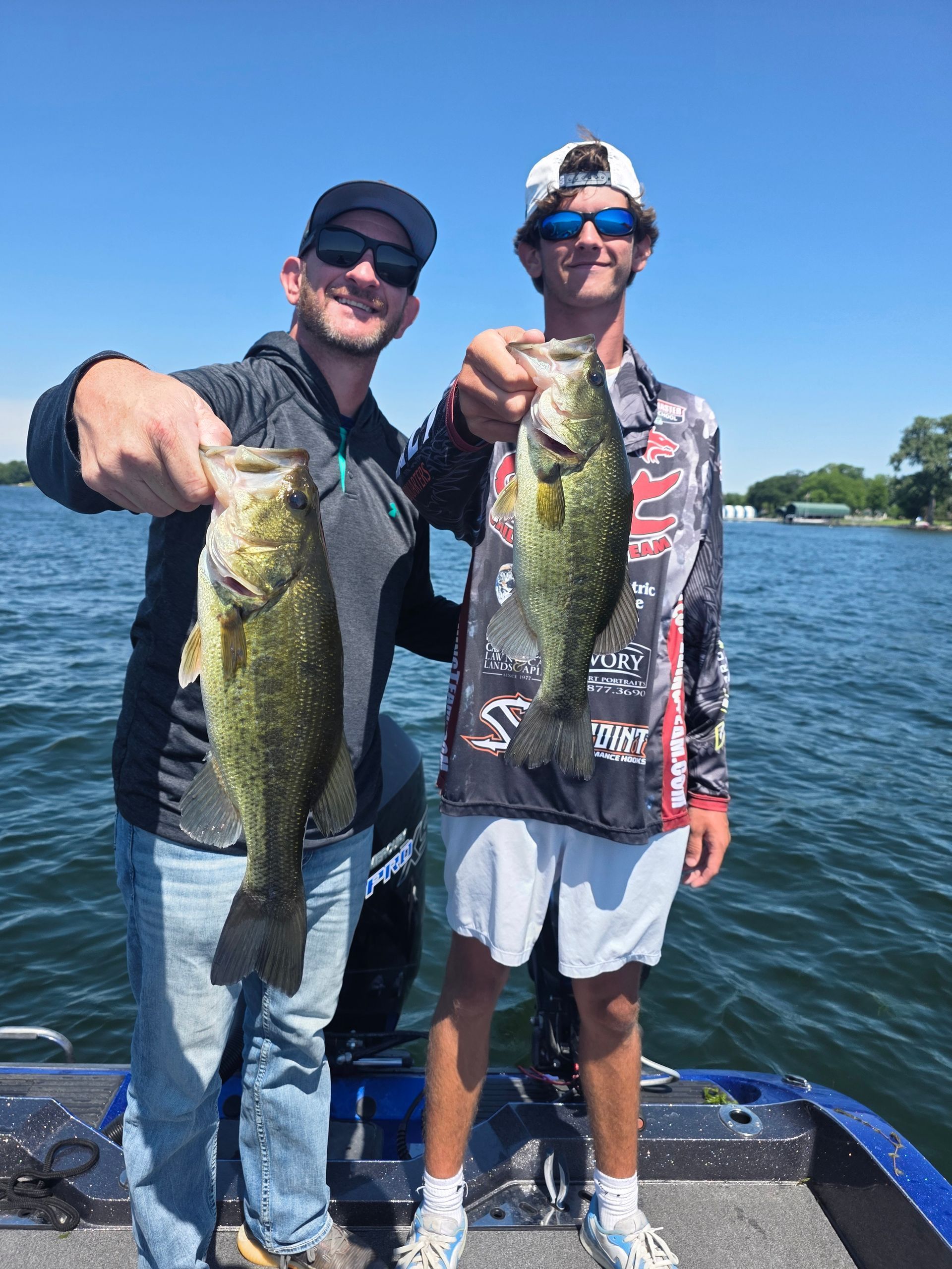 Two people on a boat hold up large fish against a sunny lake backdrop.