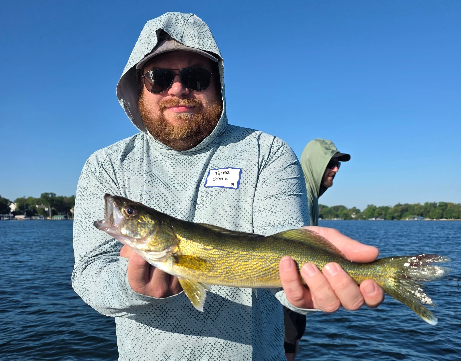 Person holding a fish on a lake, wearing a sun hat and sunglasses under a clear blue sky