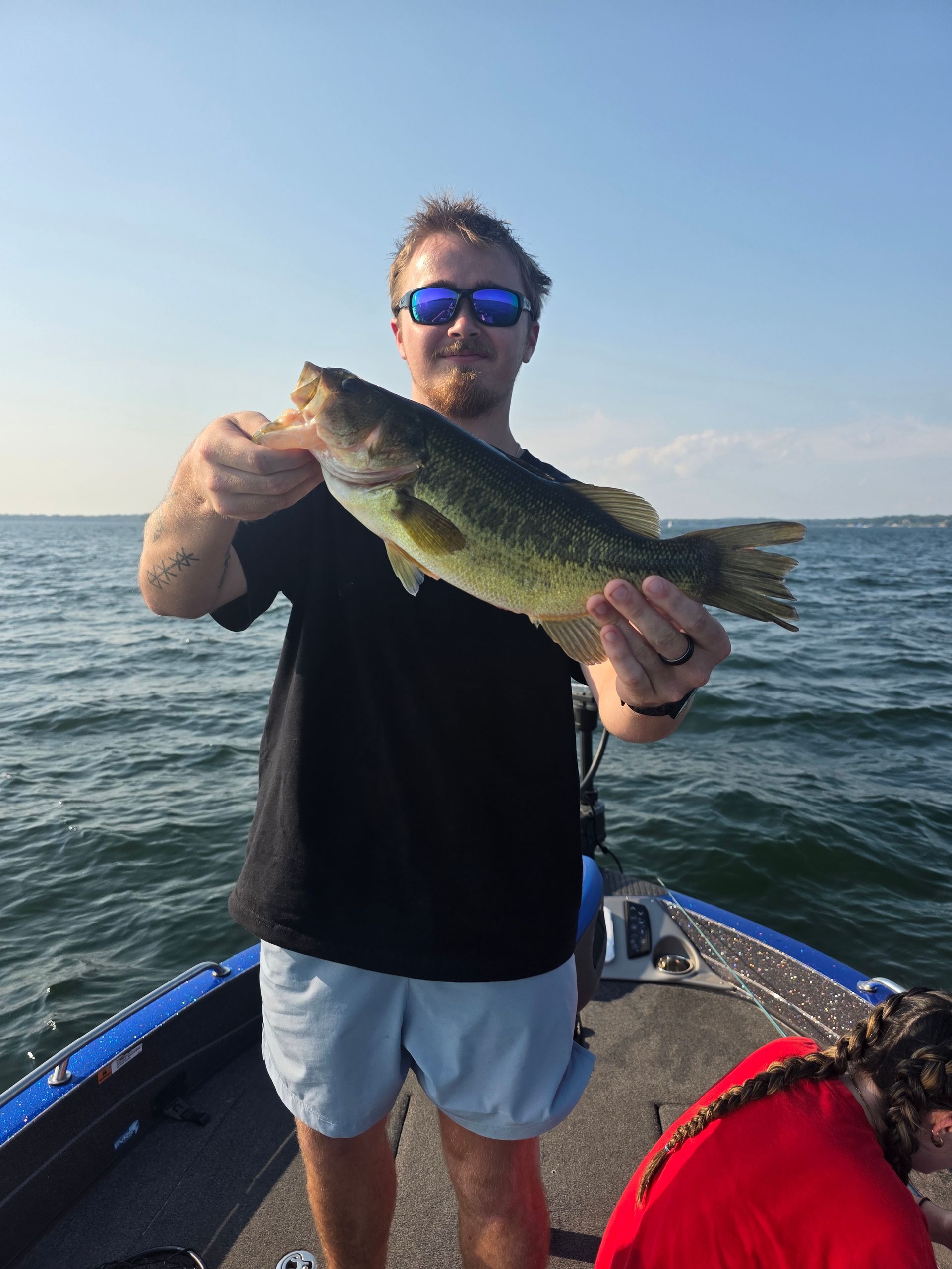 Angler holding a largemouth bass on a boat at sea, wearing sunglasses under a clear sky