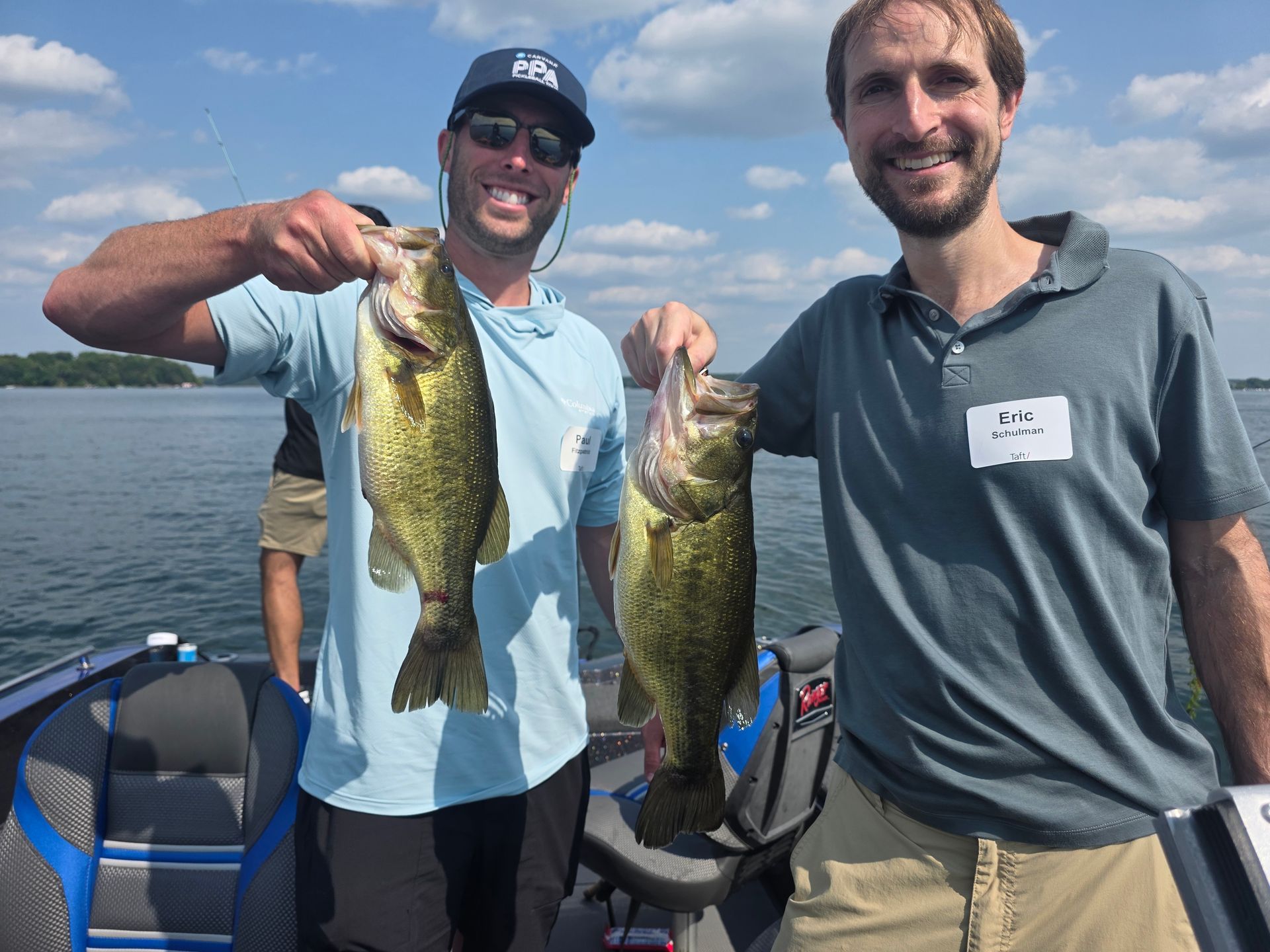 Two men on a boat holding up large fish on a sunny lake, smiling at the camera.