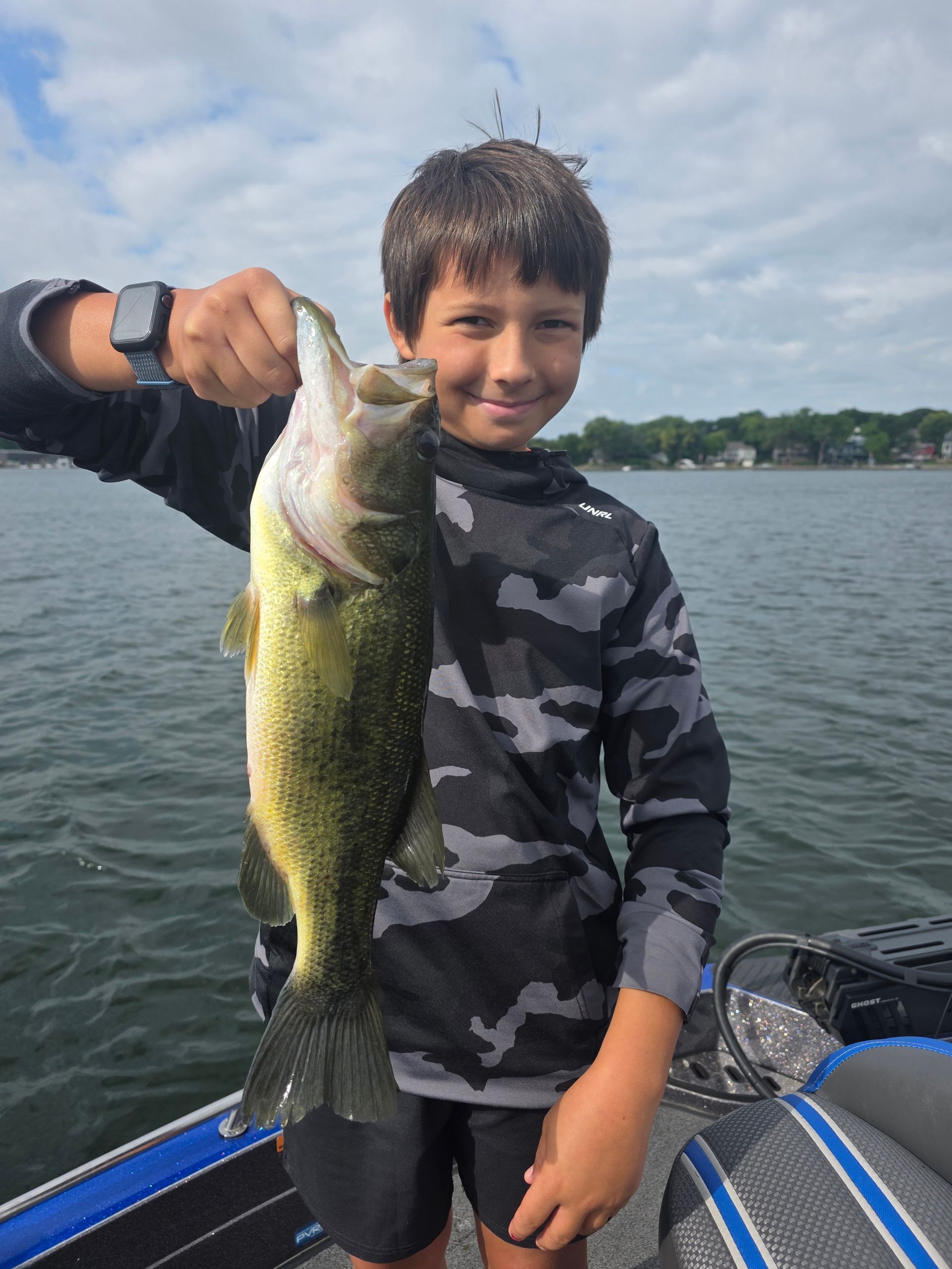 Smiling child on a boat holding a large bass over a lake, wearing a camouflage shirt.