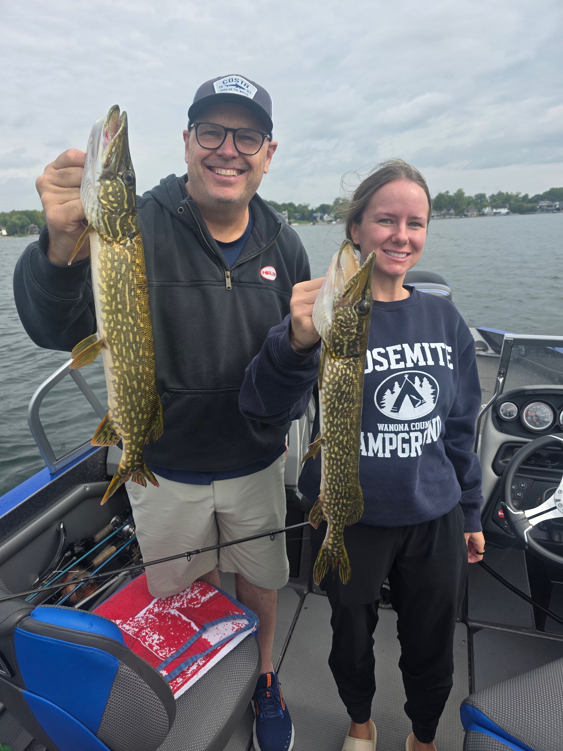 Two people on a boat proudly holding up large fish on a cloudy lake.