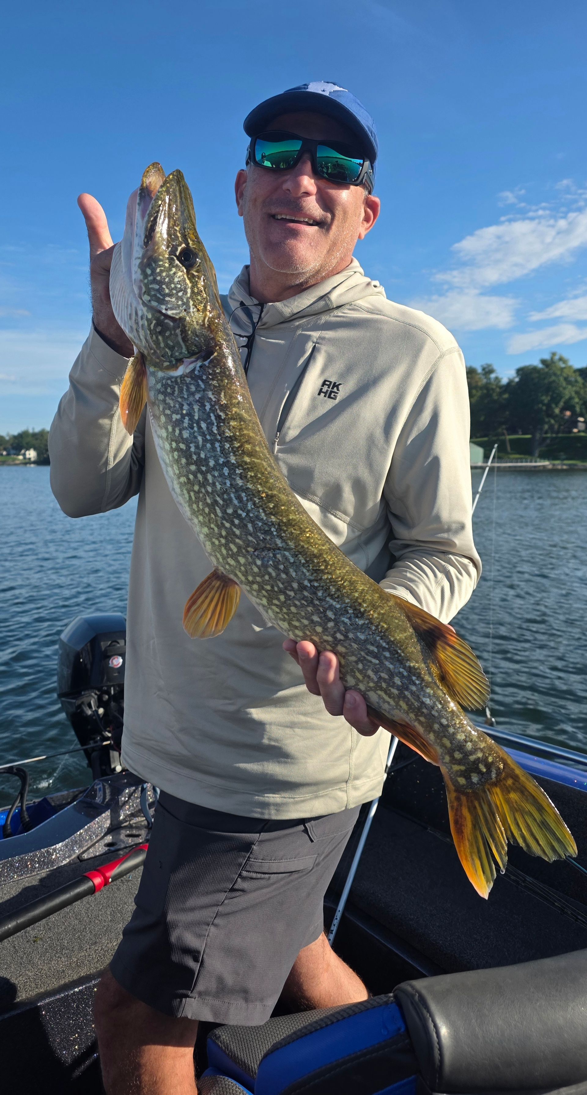 Angler holding a large northern pike on a boat, with blue sky and lake in the background