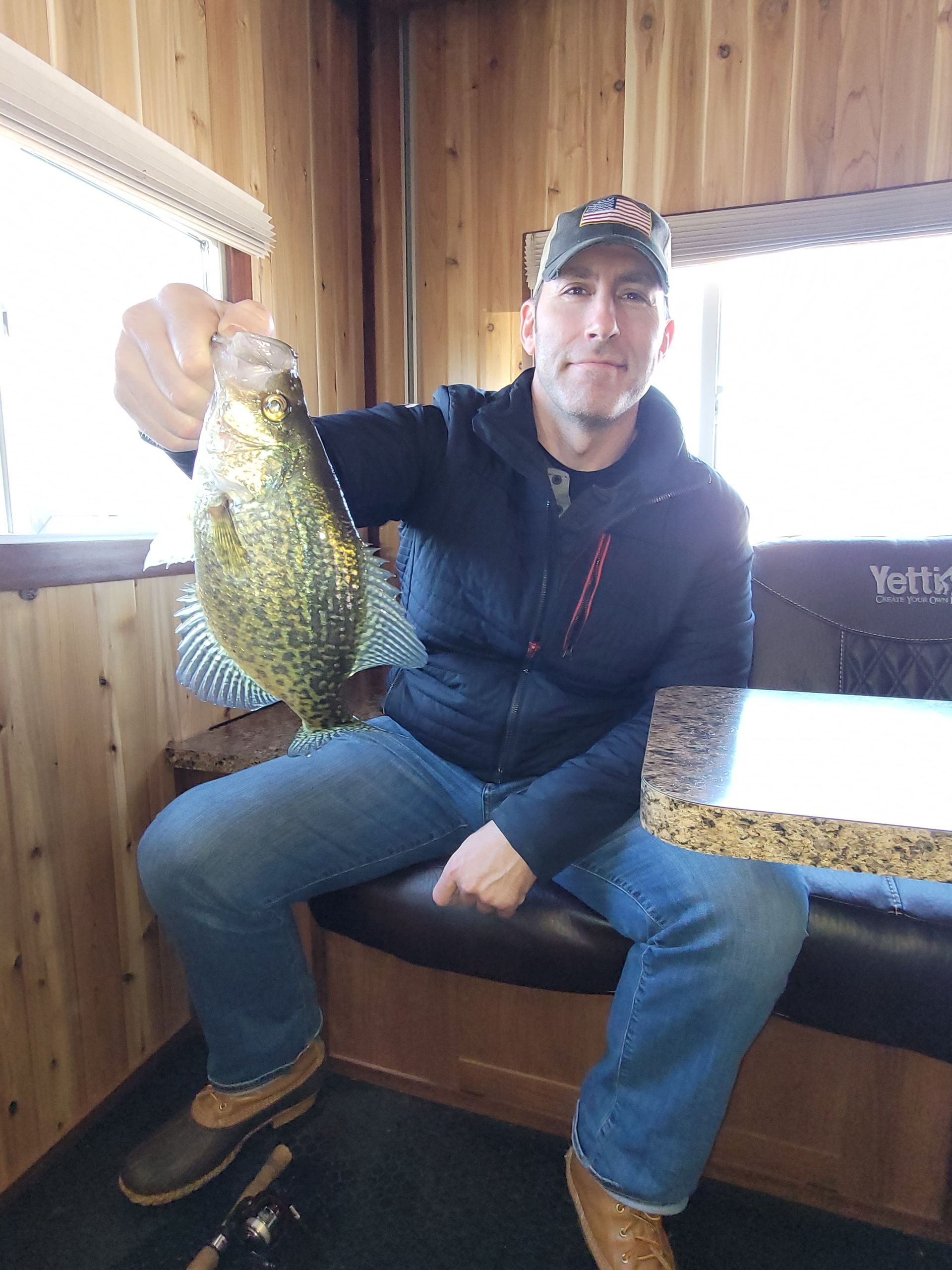 Man sitting on a bench in a wood-paneled room, holding up a large fish by a window.