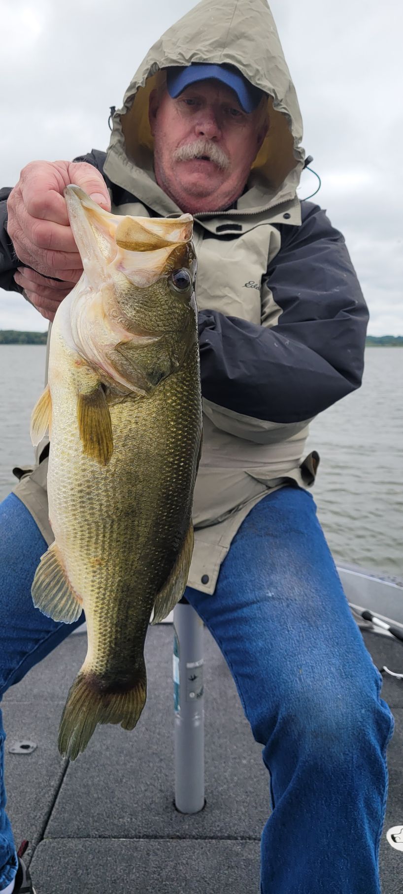 Angler in a hooded jacket holding up a large bass on a boat.