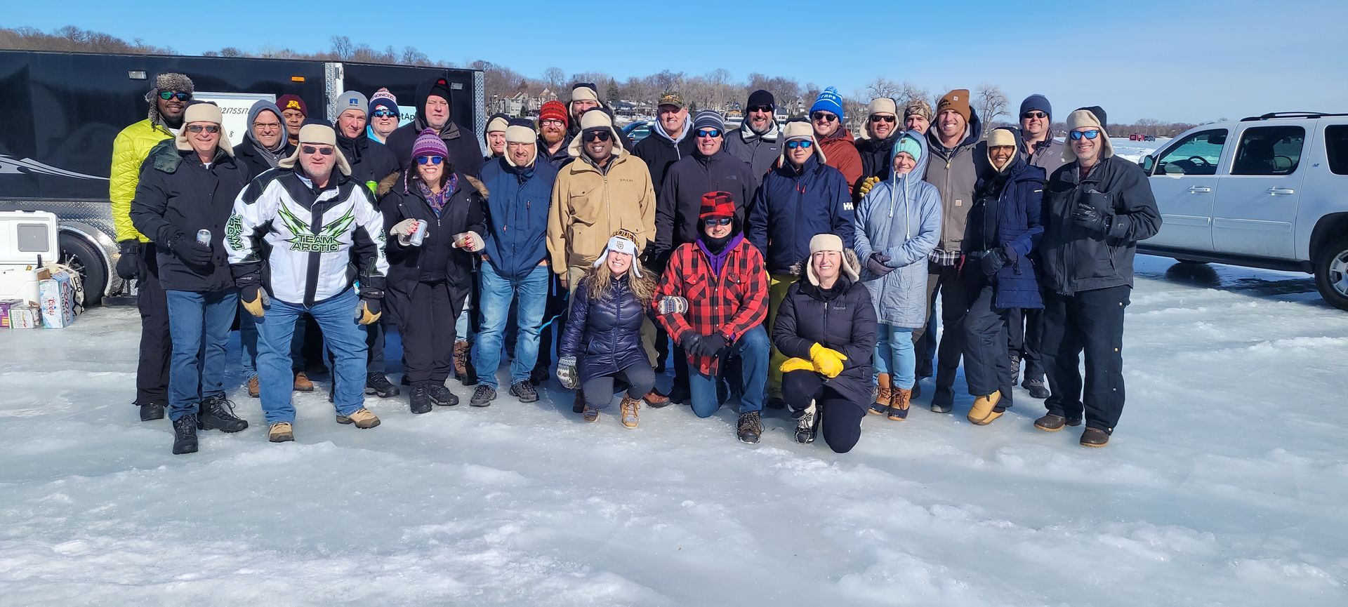 Group of people posing outdoors on snowy ice with winter gear and vehicles in the background