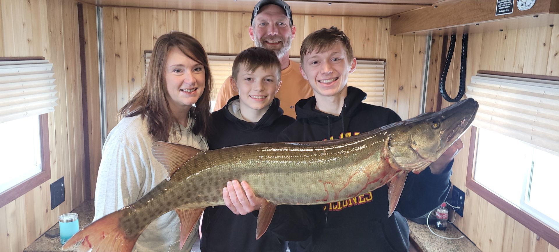 Four people indoors holding a large fish, smiling inside a wooden cabin.