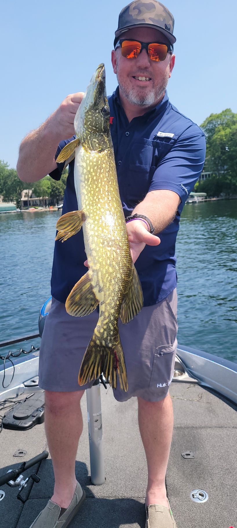 Man holding a large fish on a boat, wearing sunglasses and a cap on a sunny day.