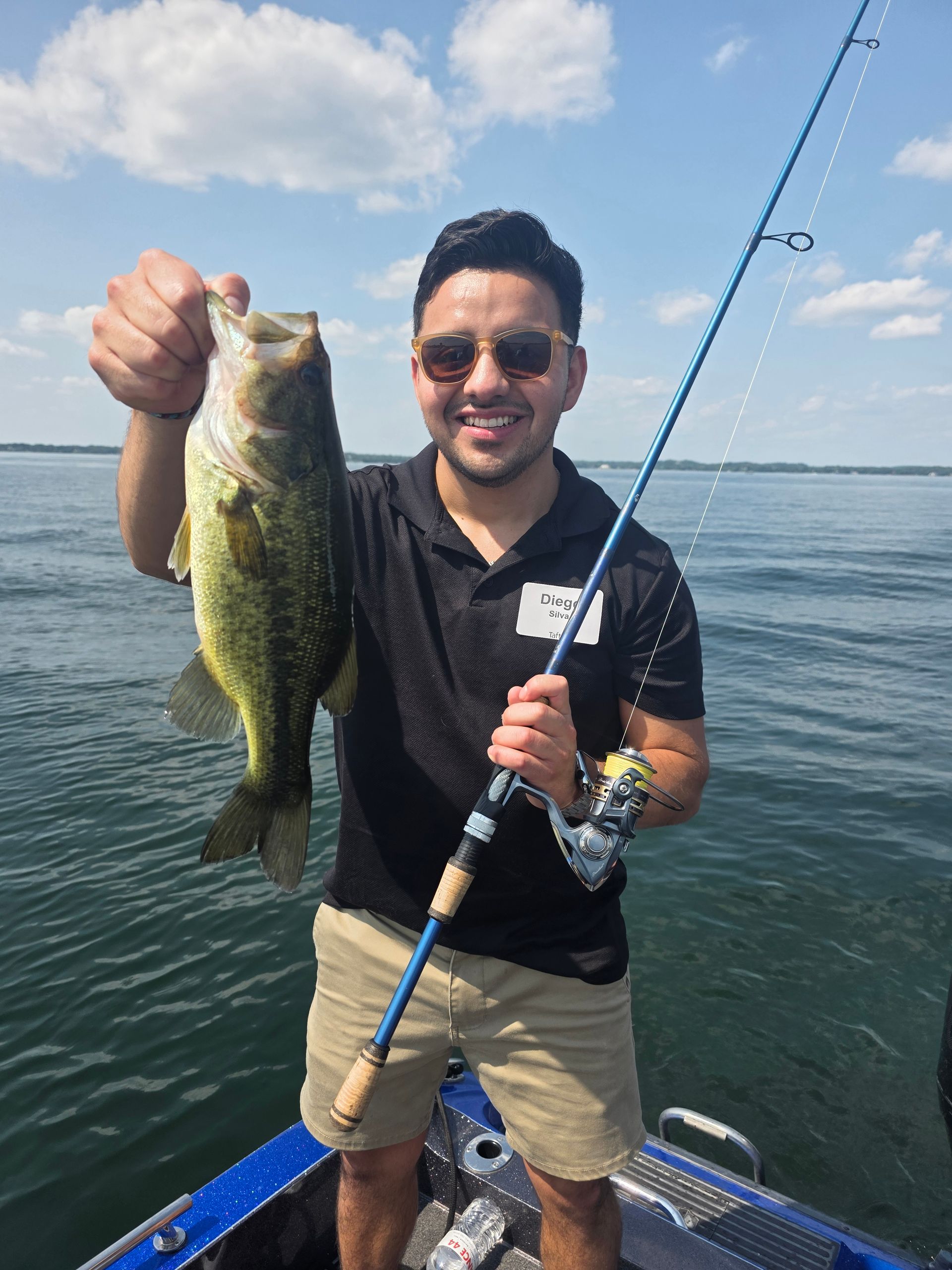 Man holding a large fish on a boat, smiling, with a fishing rod and open water behind him.