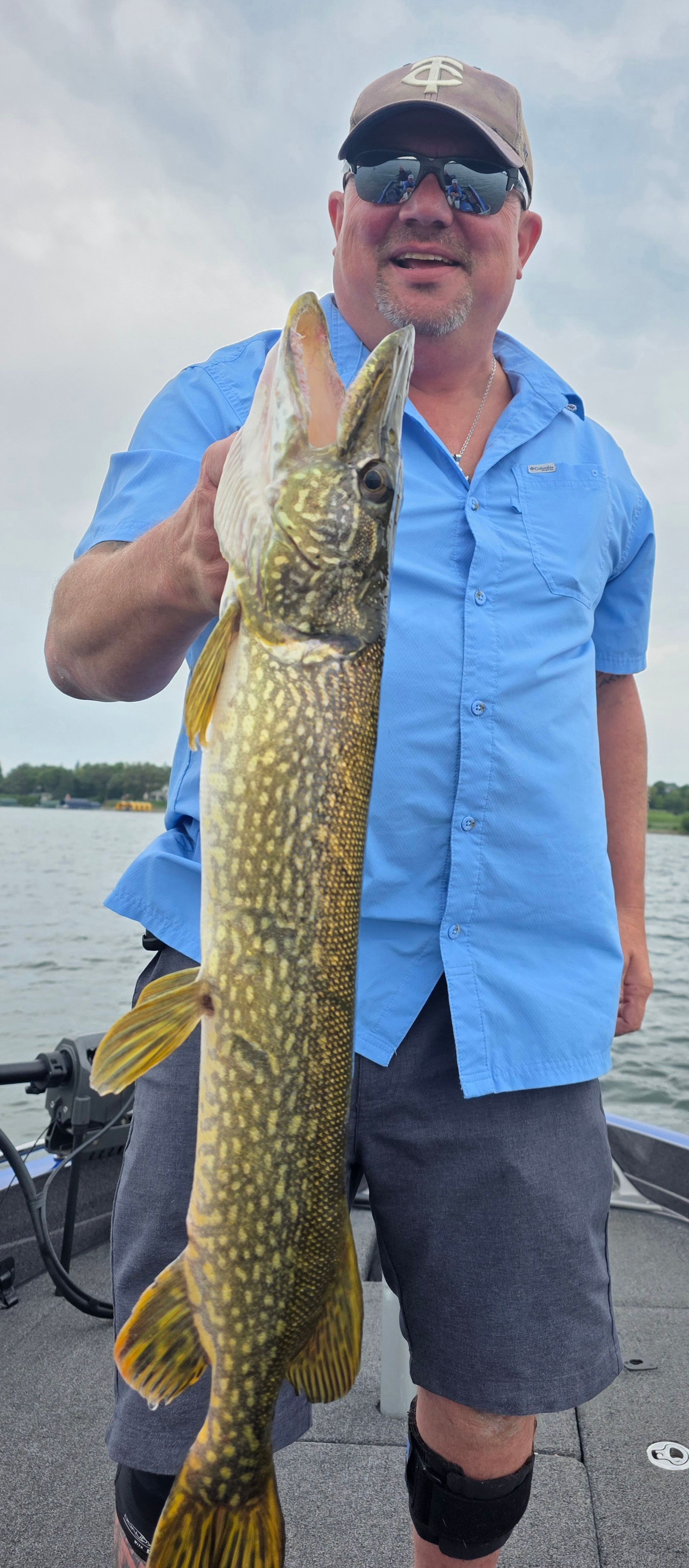 Man on a boat holding a large fish upright, wearing a blue shirt, cap, and sunglasses.