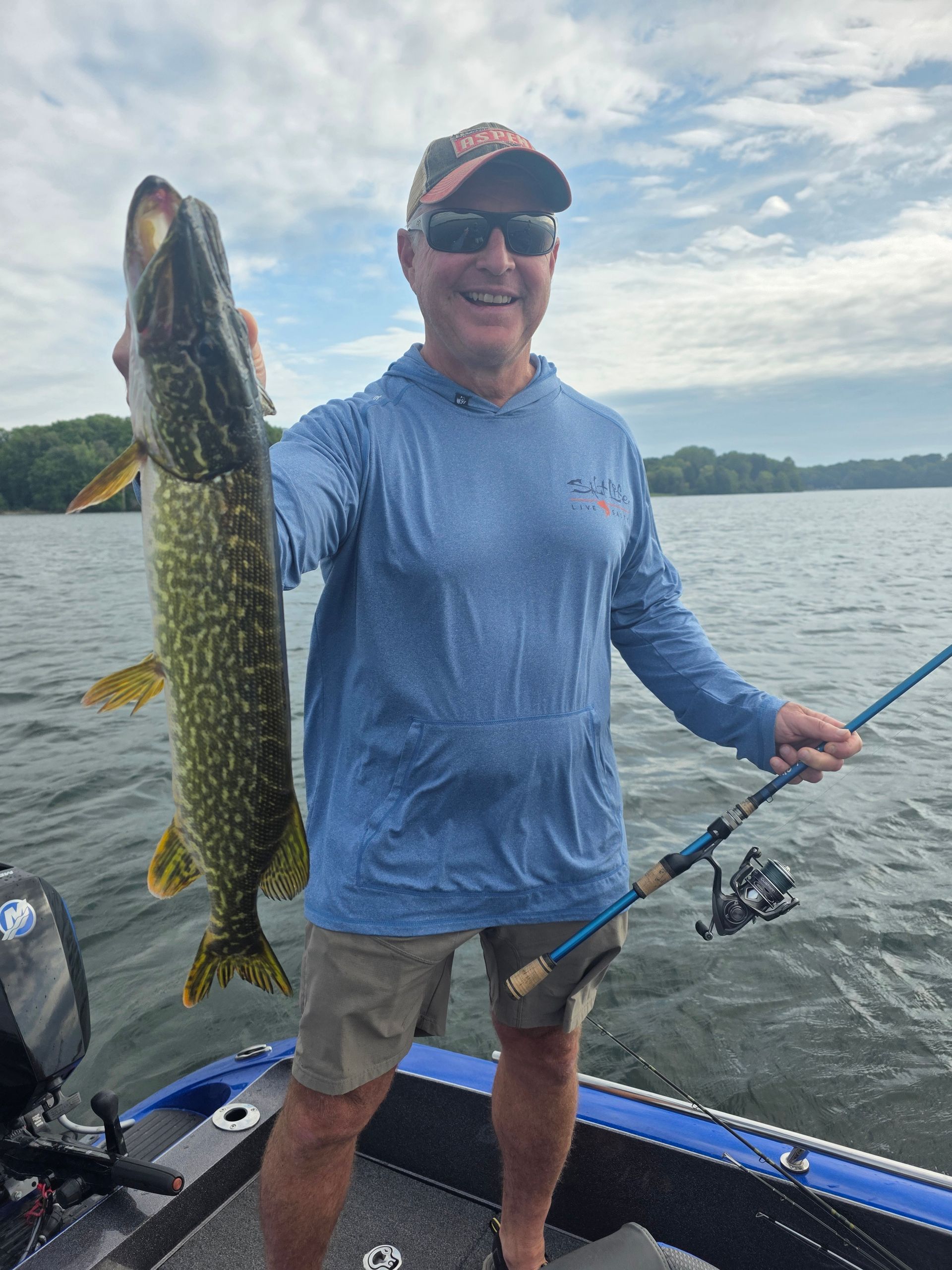 Angler on a boat holds a large fish by a lake under a cloudy sky.