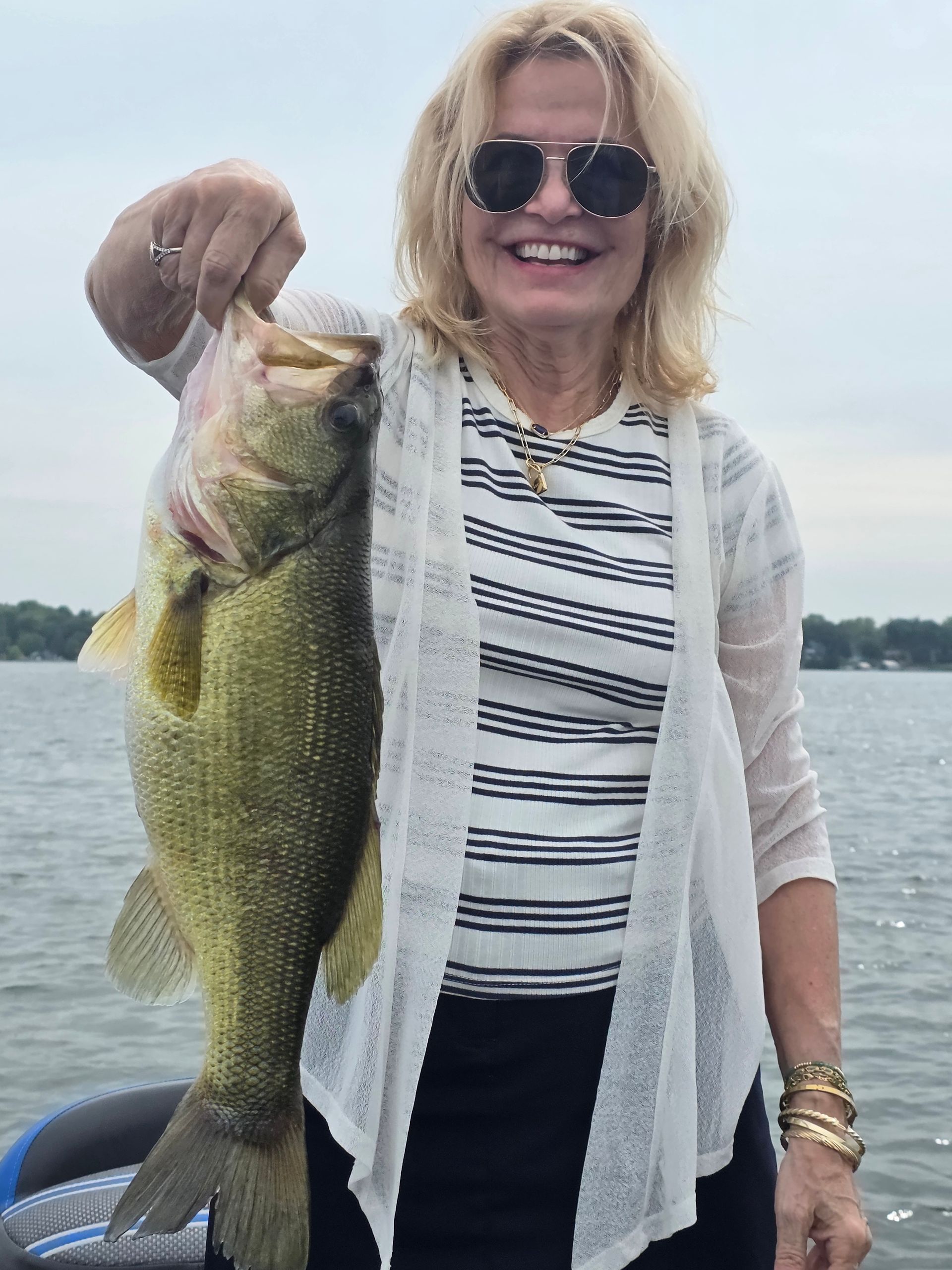 Person holding a large fish outdoors, smiling and wearing sunglasses by the water