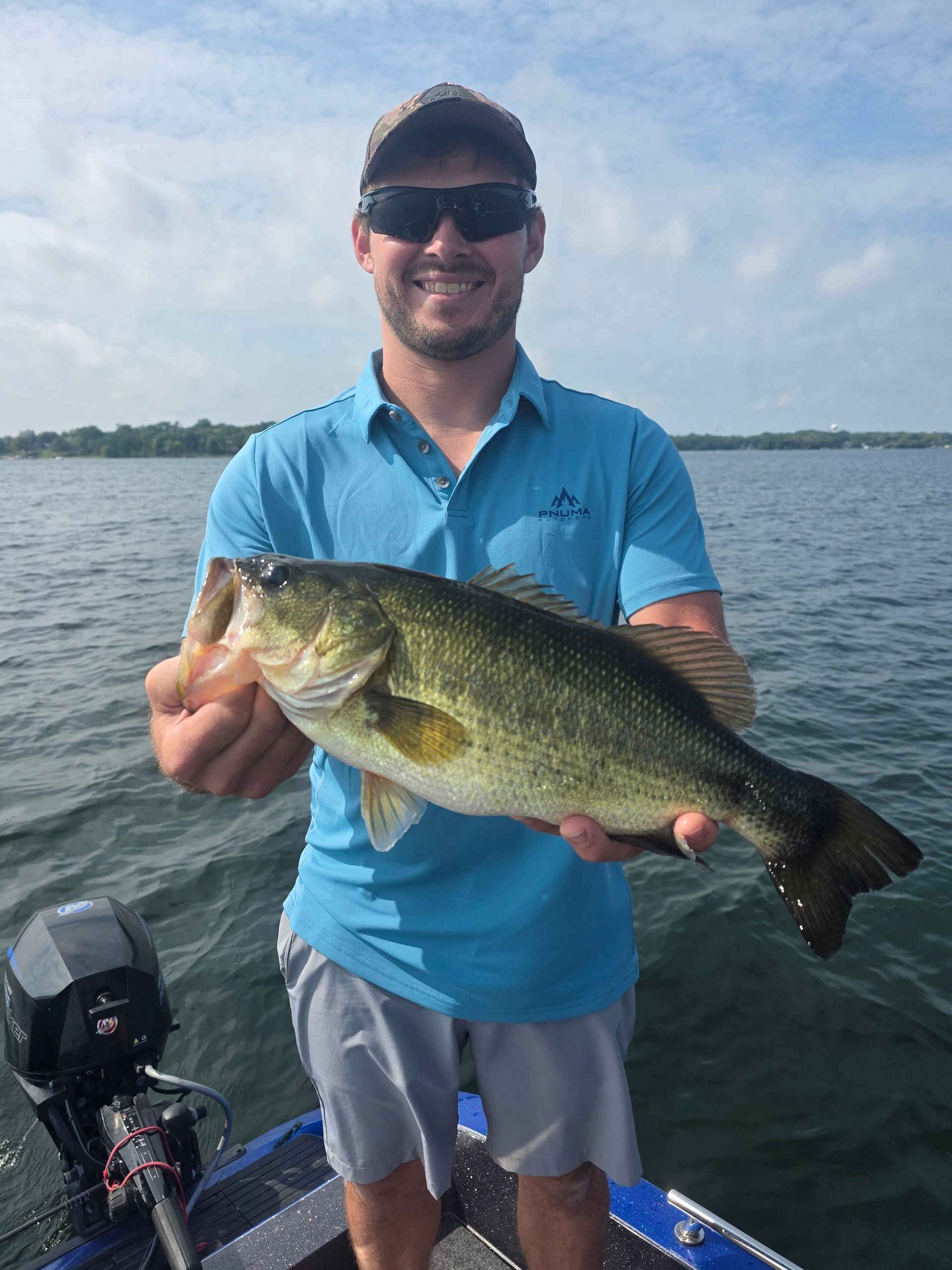 Man holding a large bass on a boat, wearing a blue shirt and sunglasses on a lake