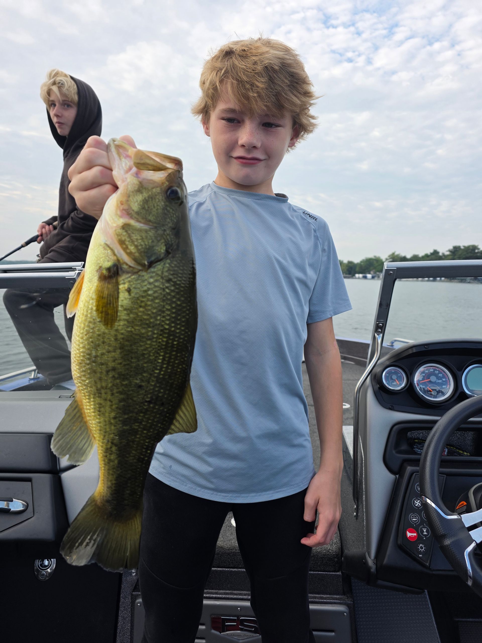 Young angler on a boat holding a large bass, with another person in a hooded jacket in the background.