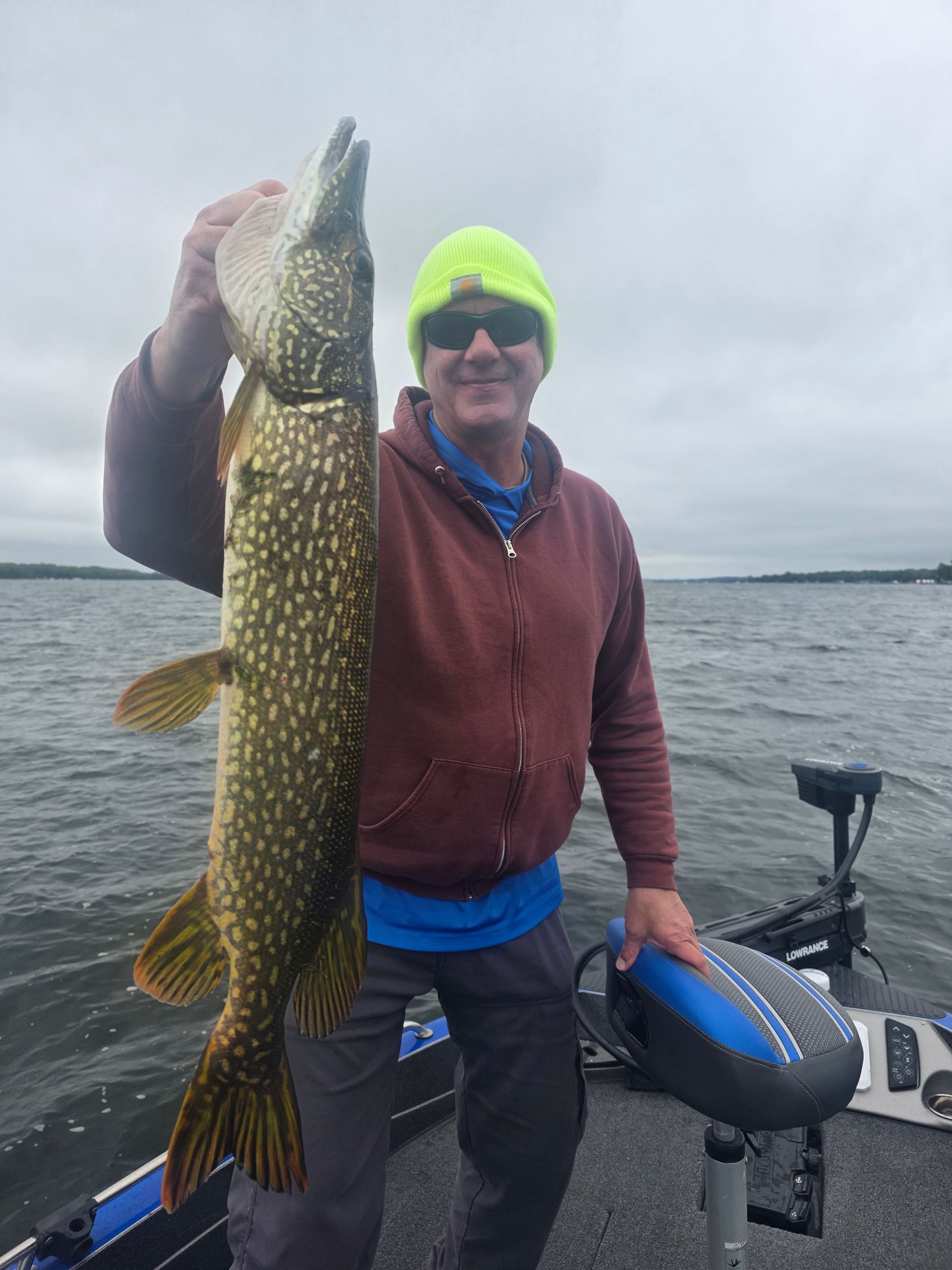 Angler on a boat holding a large northern pike by a gray lake under an overcast sky