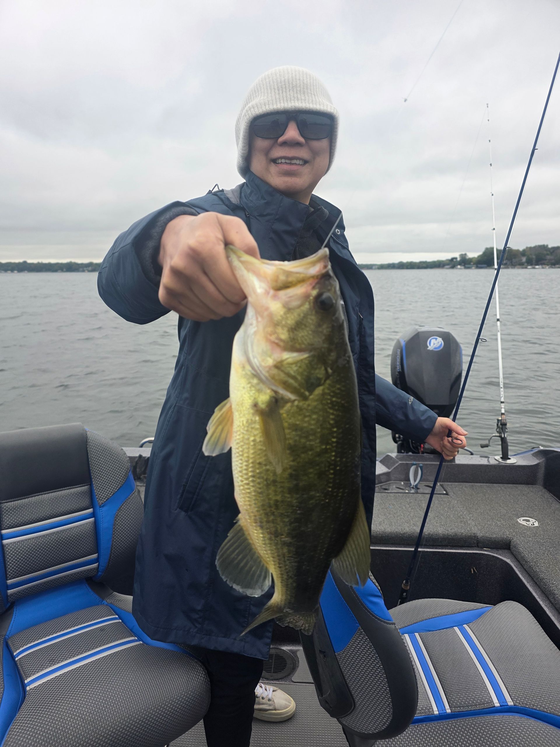 Angler on a boat holding up a large bass over a calm lake, wearing rain gear and sunglasses.