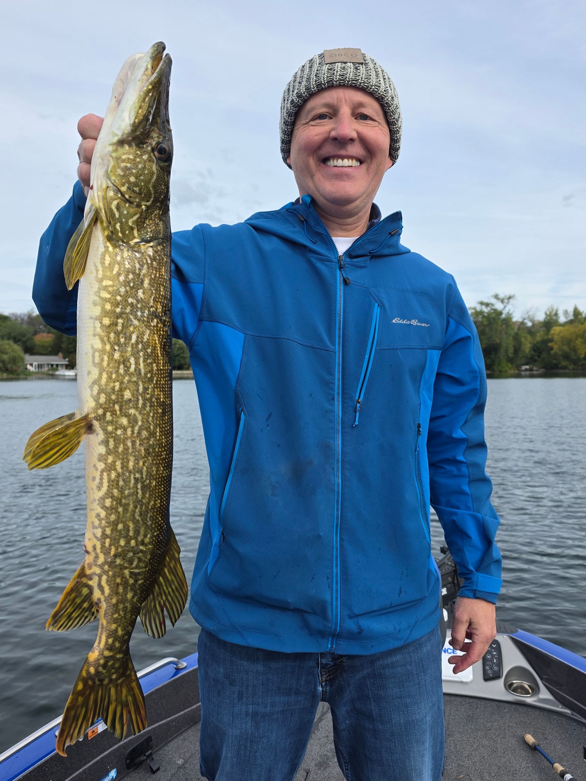 Man in blue jacket and knit cap holding up a large fish on a boat by the water