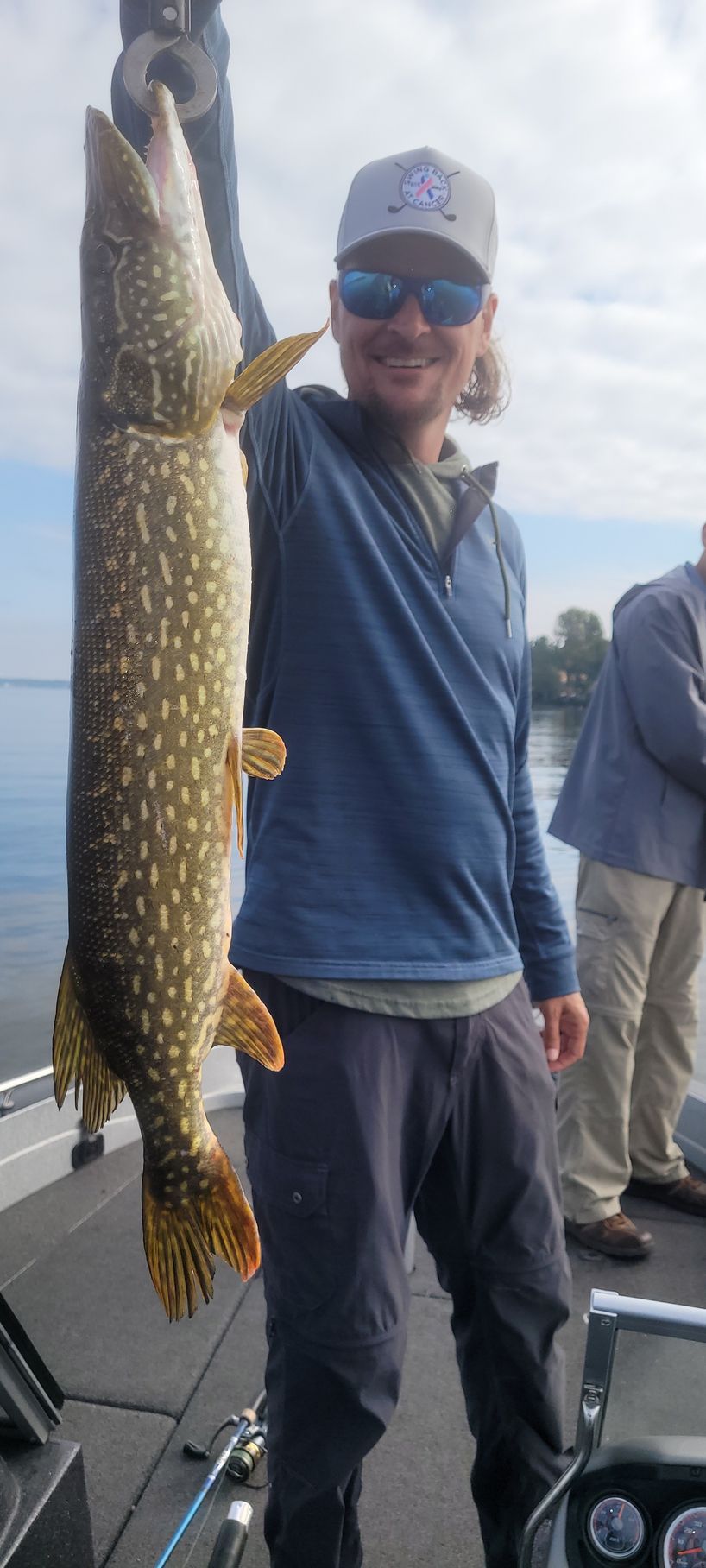 Angler holding a large northern pike on a boat, wearing a cap and sunglasses.