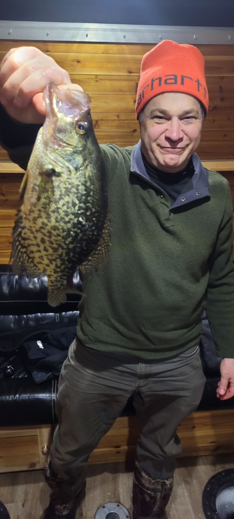 Man holding up a large mottled fish, smiling in a dark indoor setting, wearing an orange beanie and green jacket