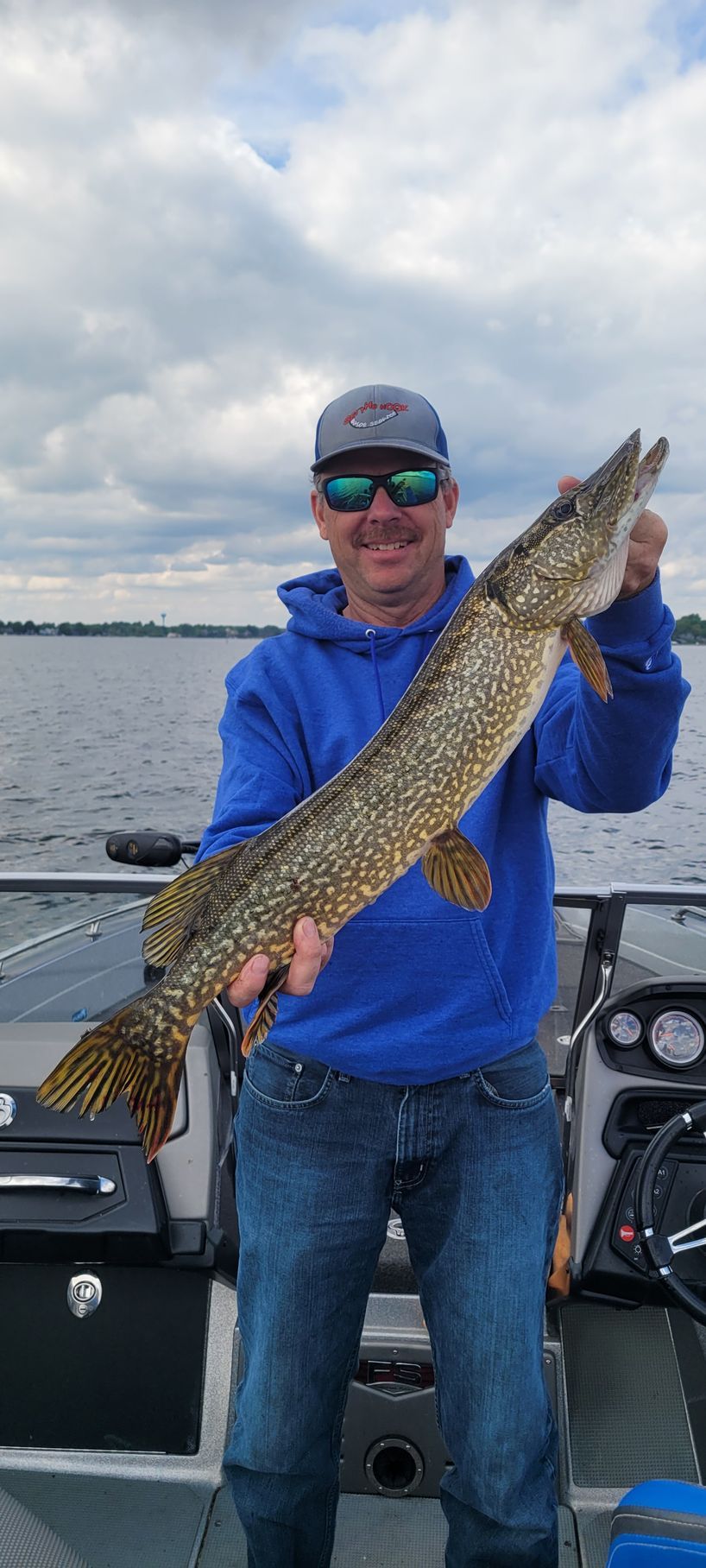 Smiling angler on a boat holding a large fish against a cloudy lake backdrop