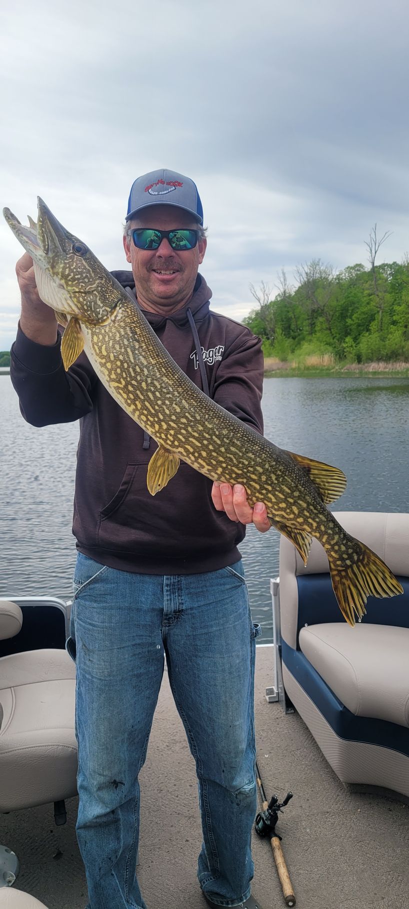 Man holding a large fish on a boat, wearing sunglasses and a cap on a cloudy day
