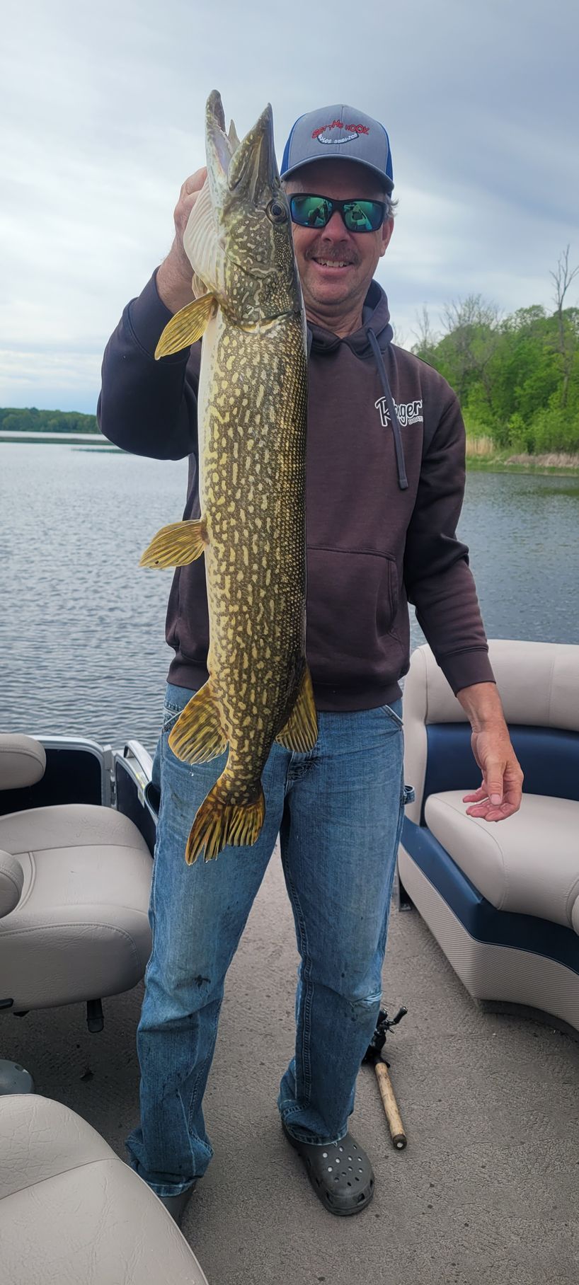 Man holding a large northern pike on a boat beside a lake