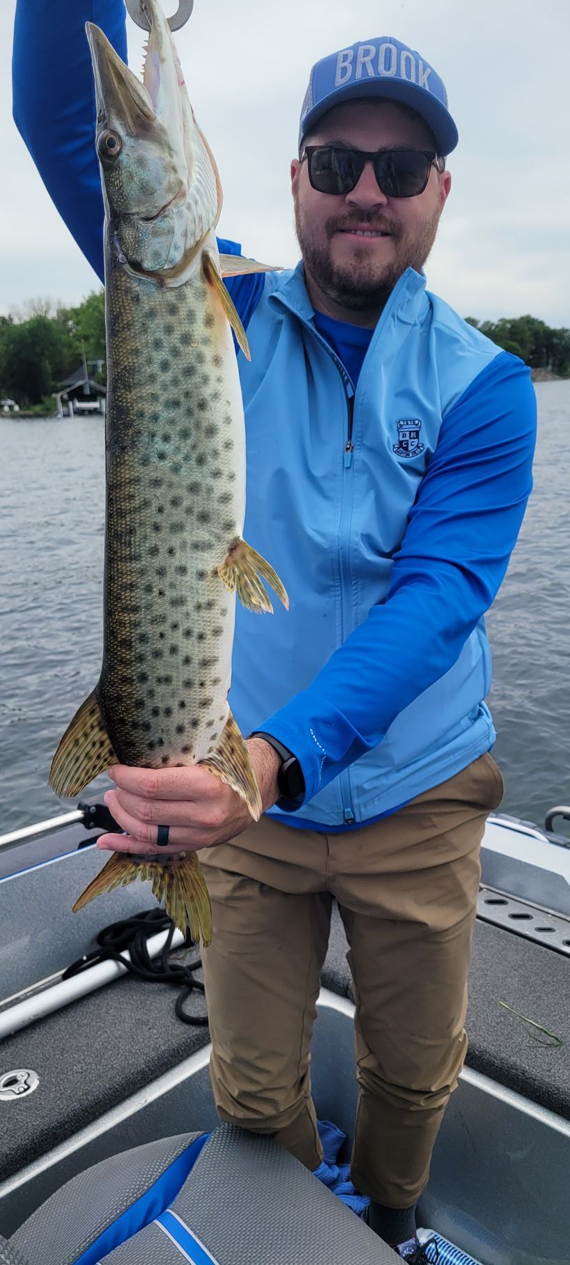 Man in blue shirt holding a large spotted fish on a boat docked on calm water