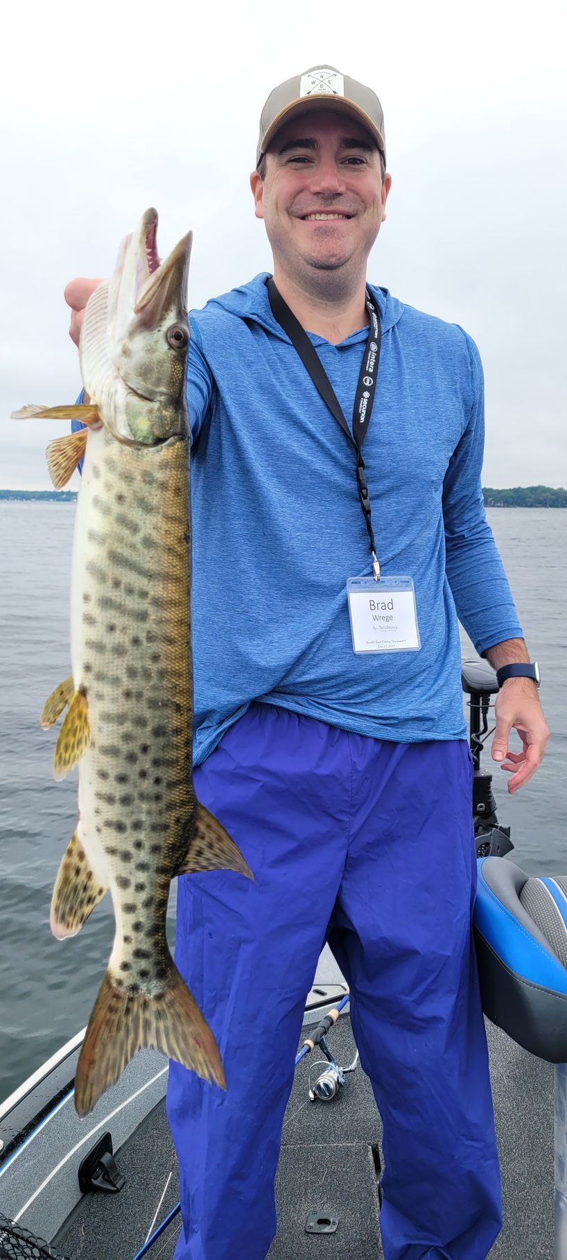 Angler on a boat holding a large fish over the water, wearing a blue shirt and pants.