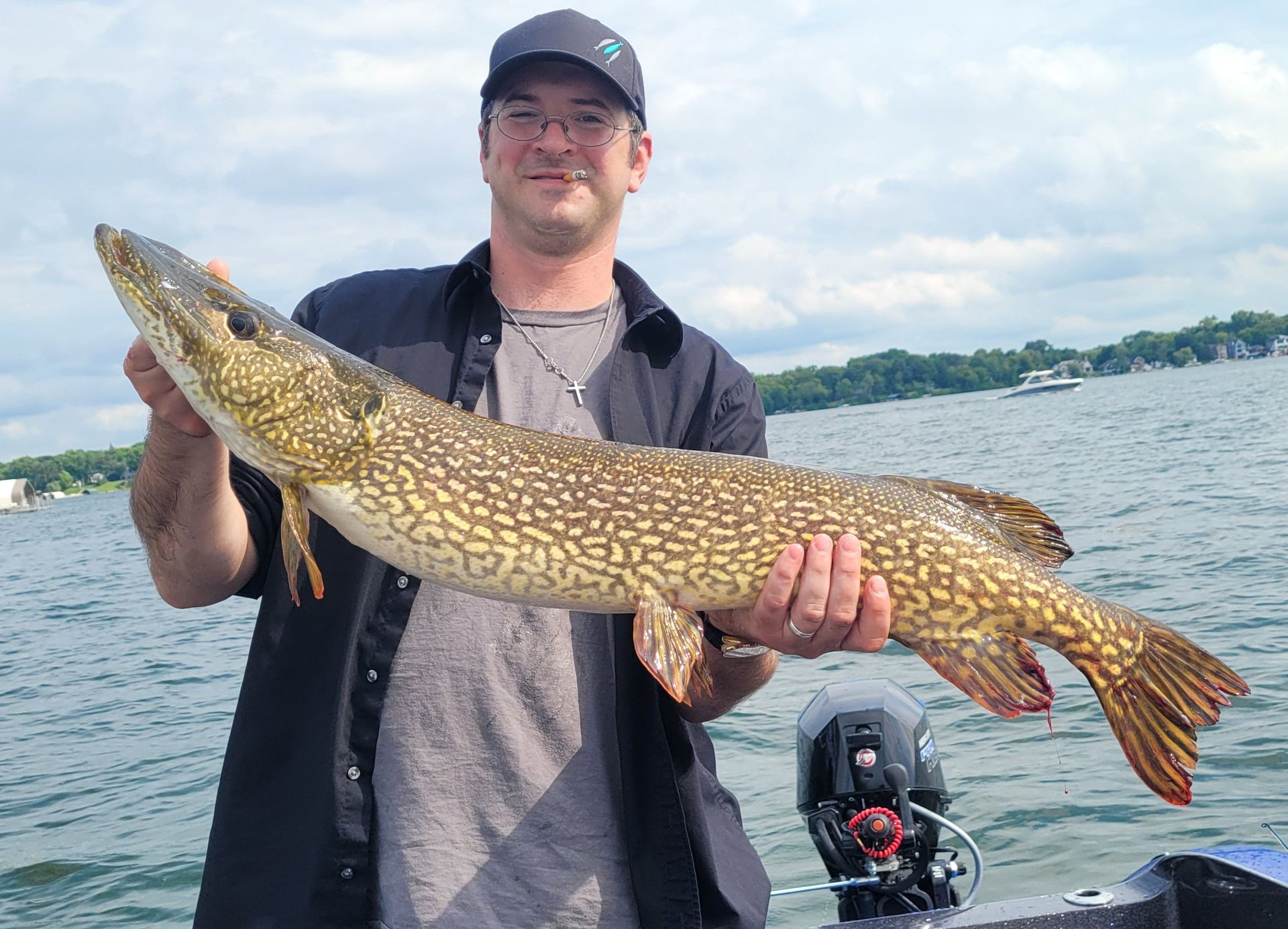 Person holding a large northern pike on a boat with water and shoreline in the background