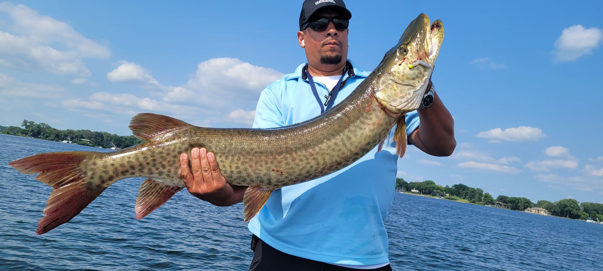 Man holding a large fish on a boat, wearing a blue shirt and cap on a sunny lake