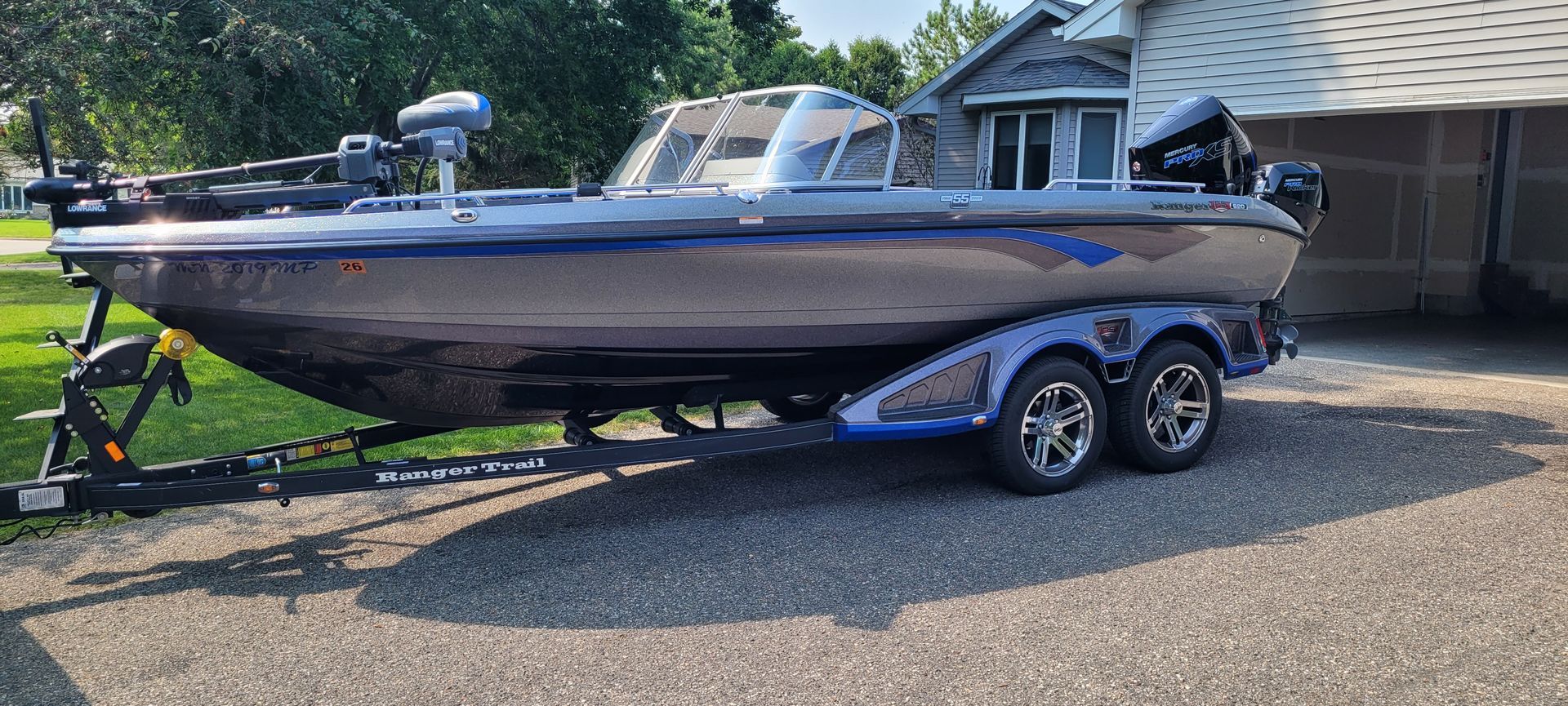 Bass boat on a trailer in a driveway, with a blue cover and windshield under bright sun