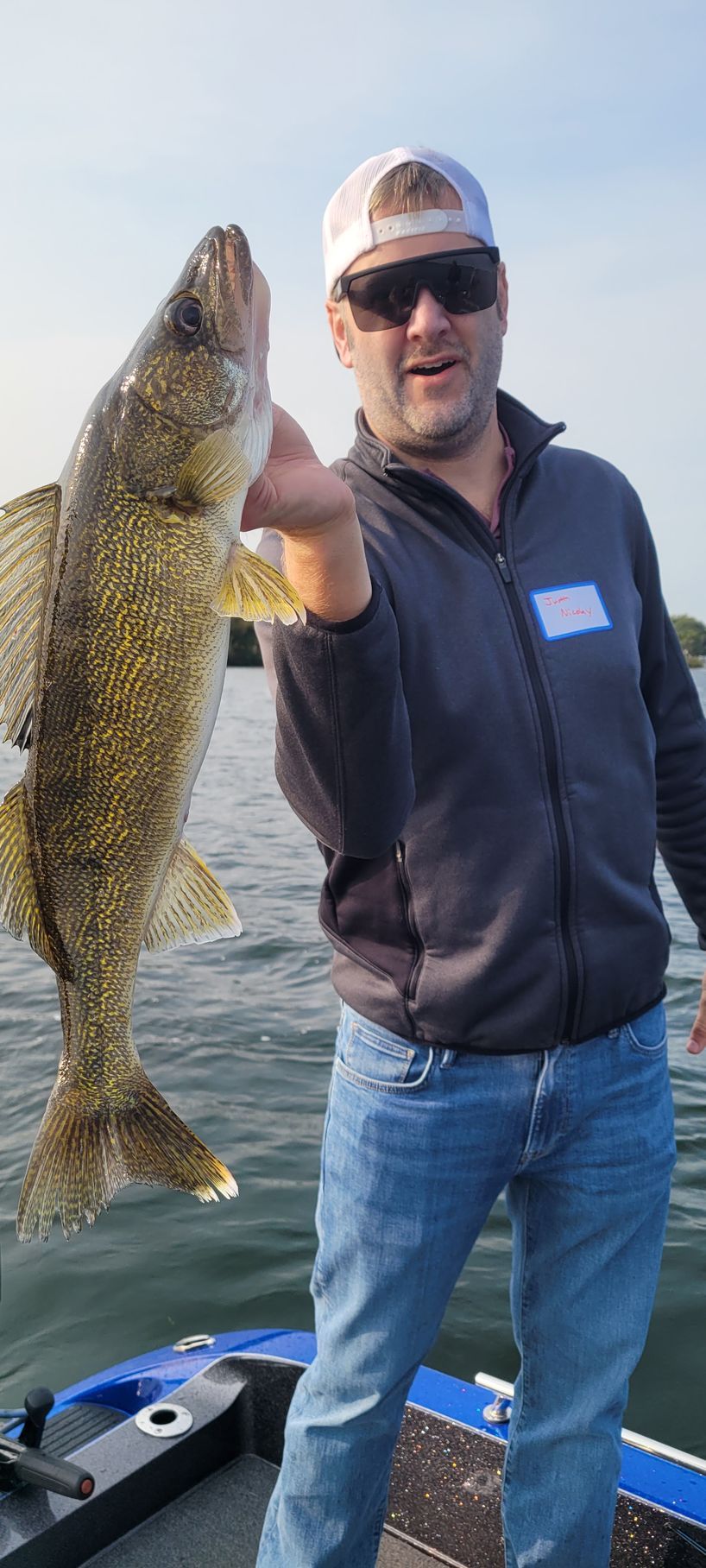 Angler on a boat holding up a large fish over the water, wearing sunglasses and a cap.