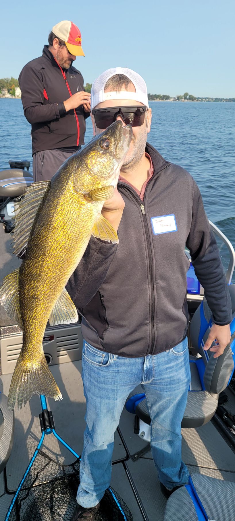 Man holding a large fish on a boat, with another person fishing in the background on the water.