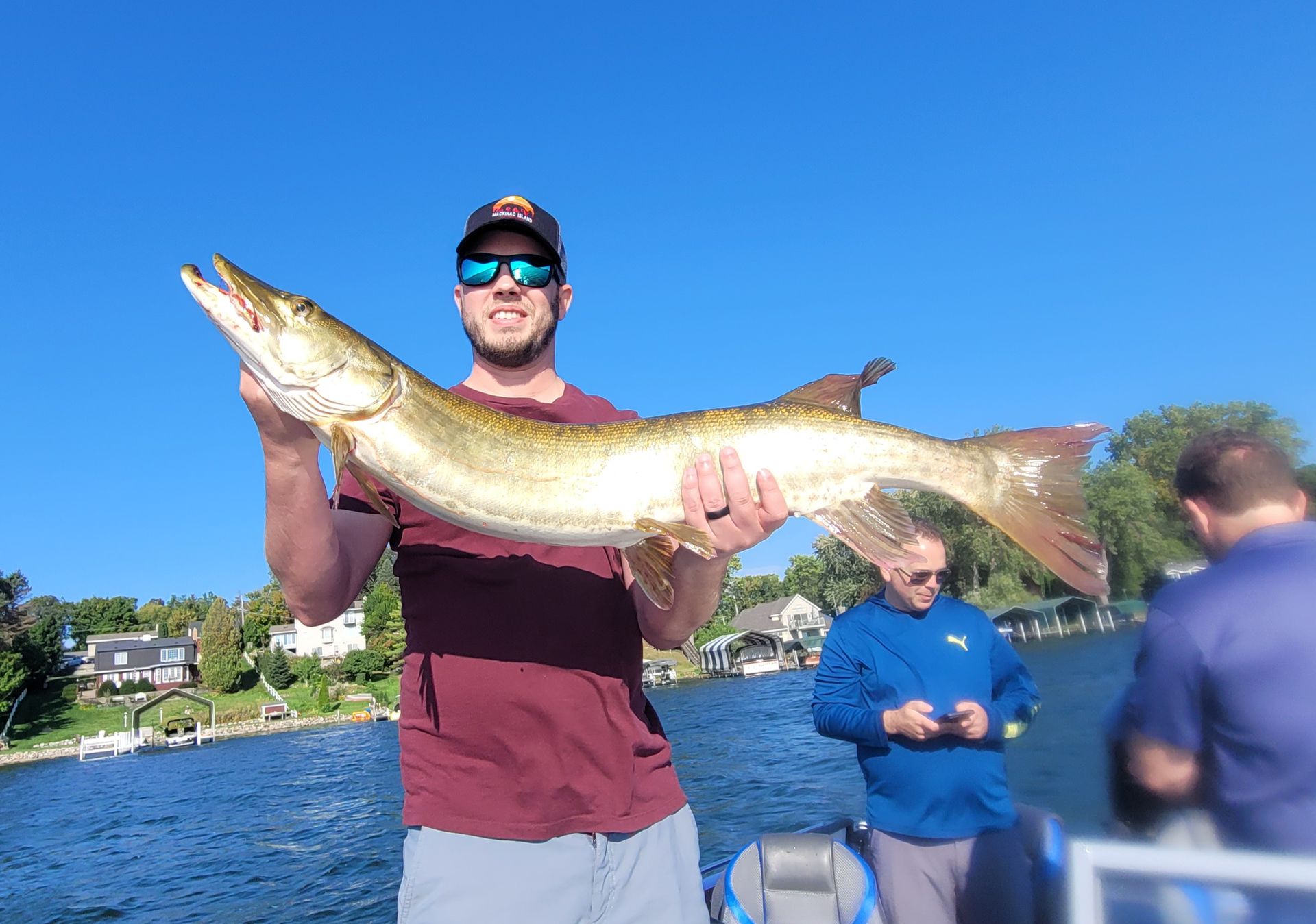 Man holding a large fish on a boat with blue water and trees in the background