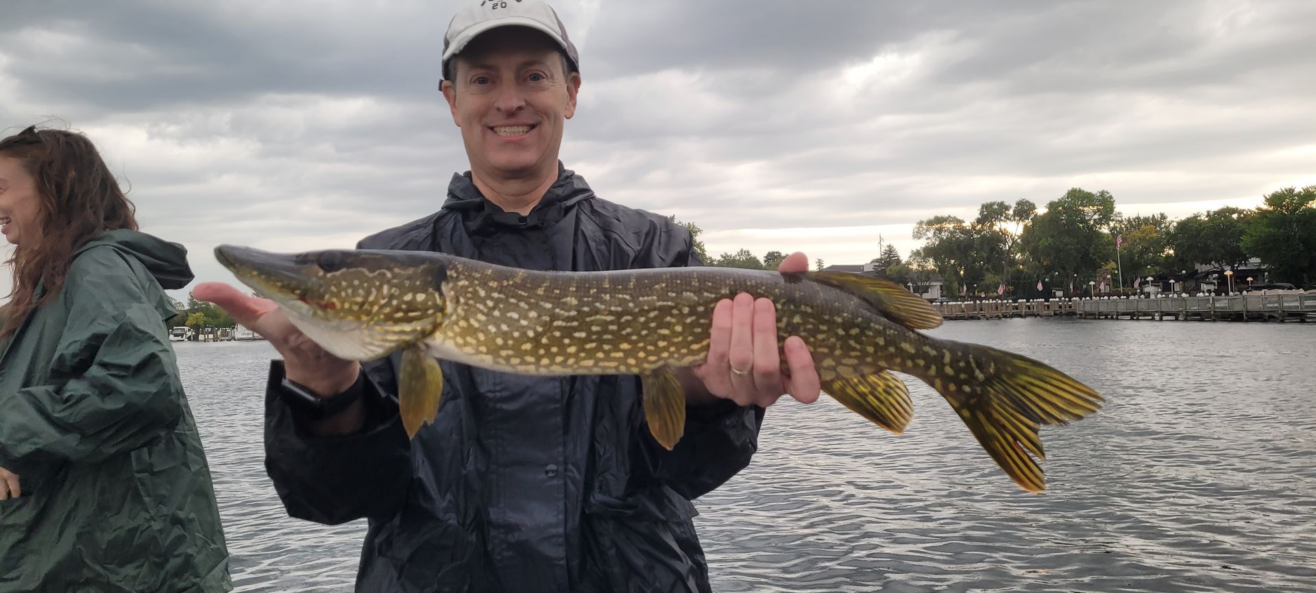 Person holding a large fish by the water at dusk, wearing a cap and jacket