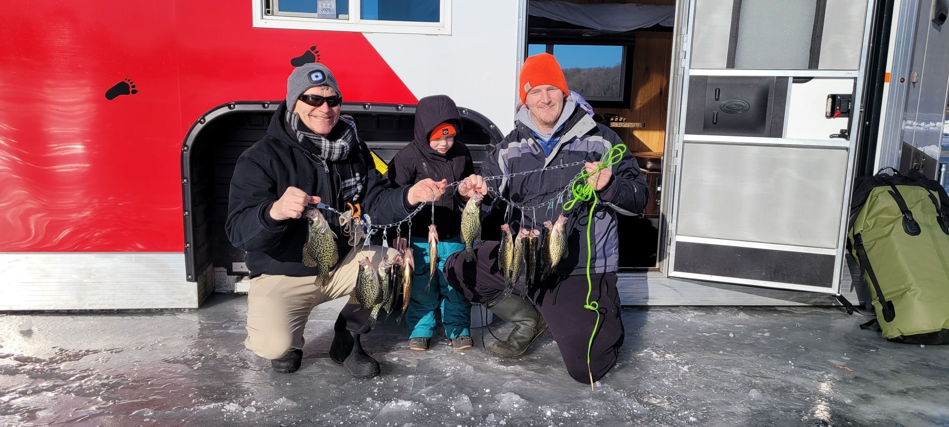 Three anglers kneel on ice holding freshly caught fish beside a red building.