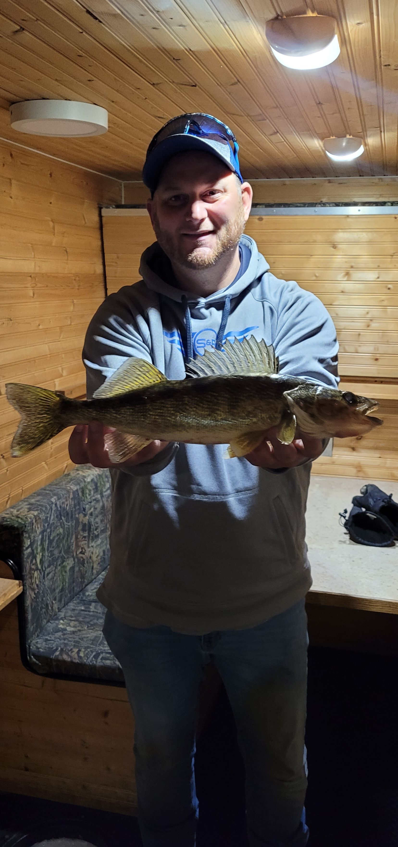 Man holding a large fish indoors in a wood-paneled room, wearing a cap and jacket.