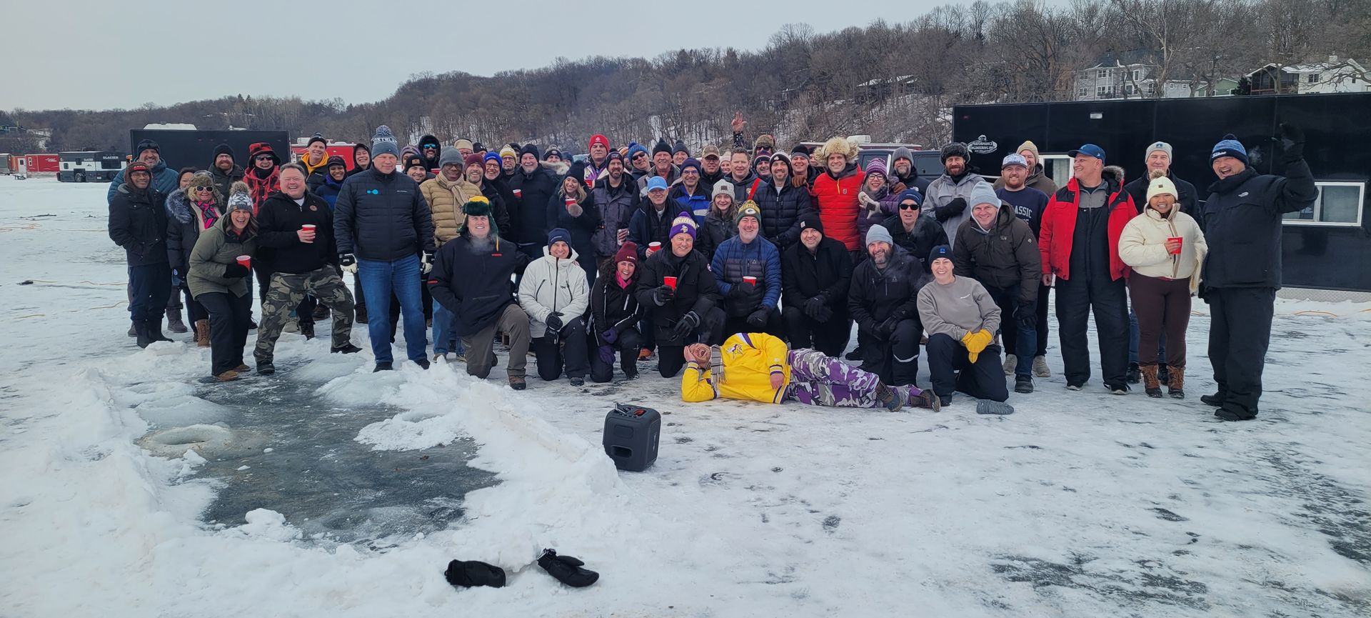 Large group posing on a snowy frozen lake with ice-fishing shelters and winter trees in the background.