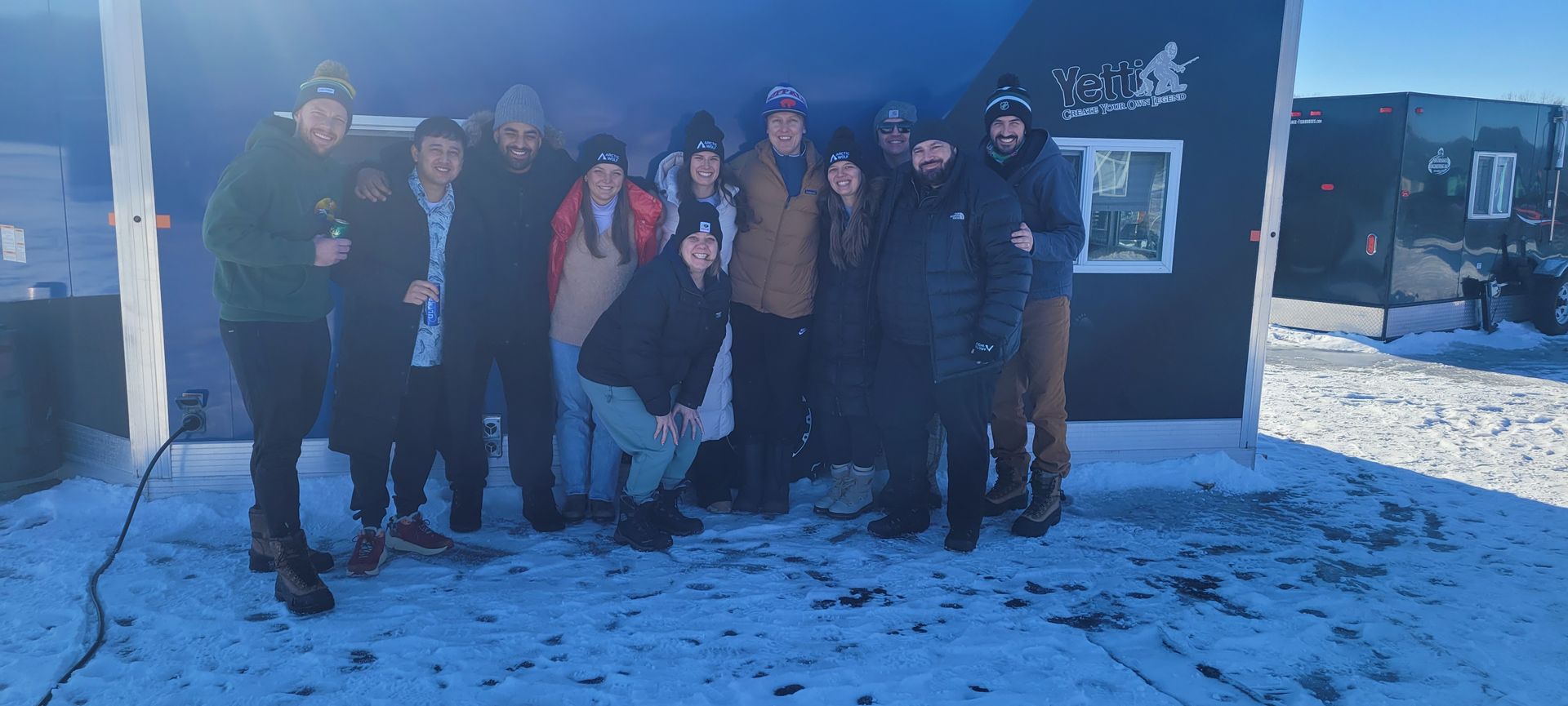 Group of people posing outside a building in the snow, wearing winter coats and standing in sunlight.