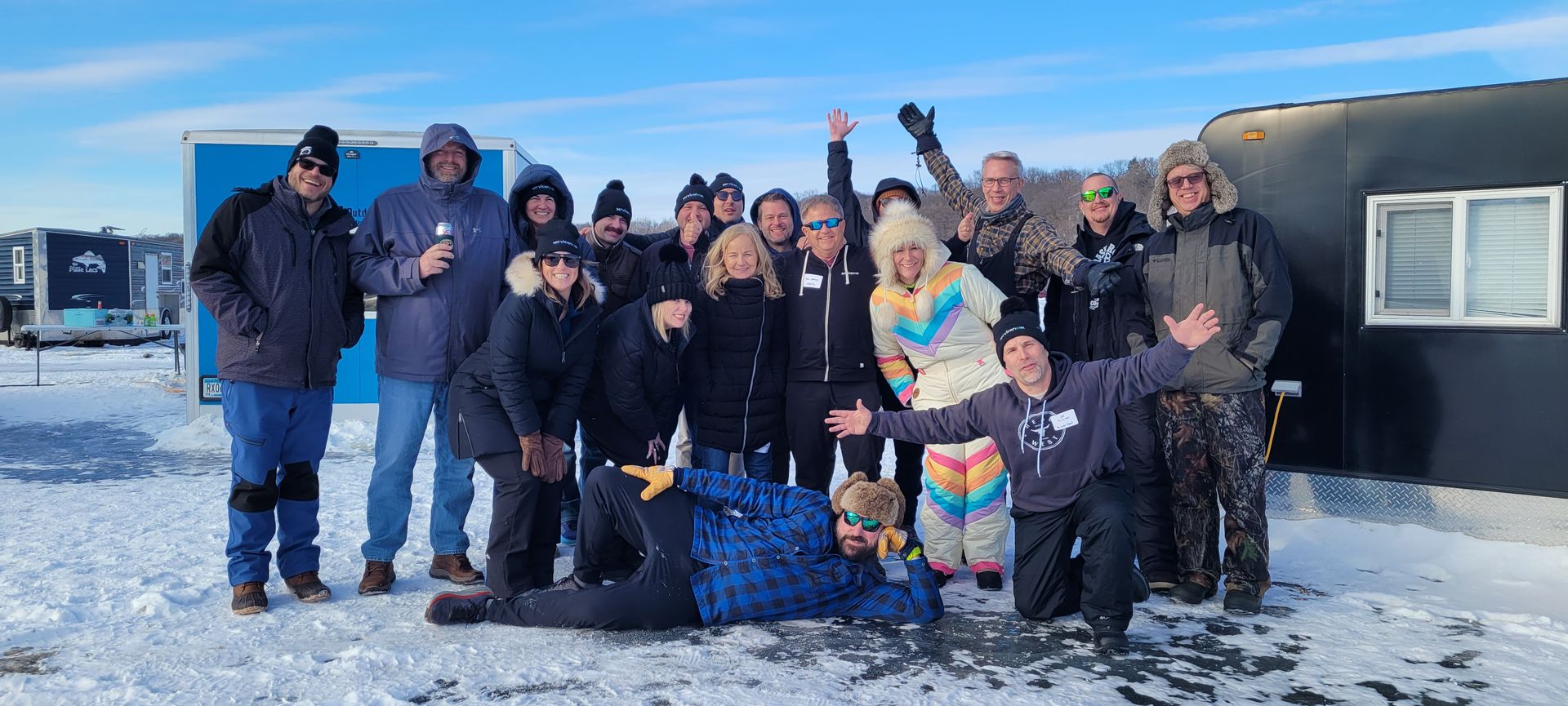 Group of people posing in winter clothes beside a dark bus or trailer on a snowy shoreline under a blue sky