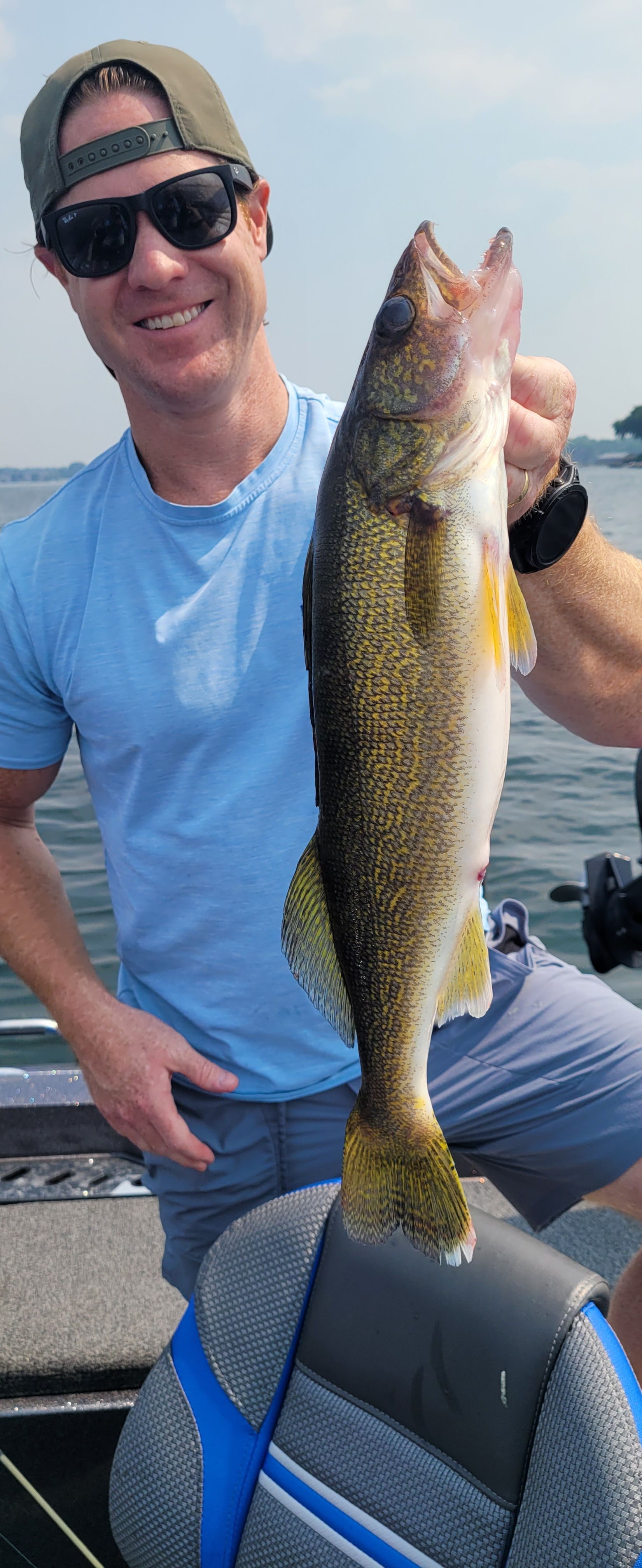 Man holding a large fish by a boat seat on the water, wearing sunglasses and a cap.