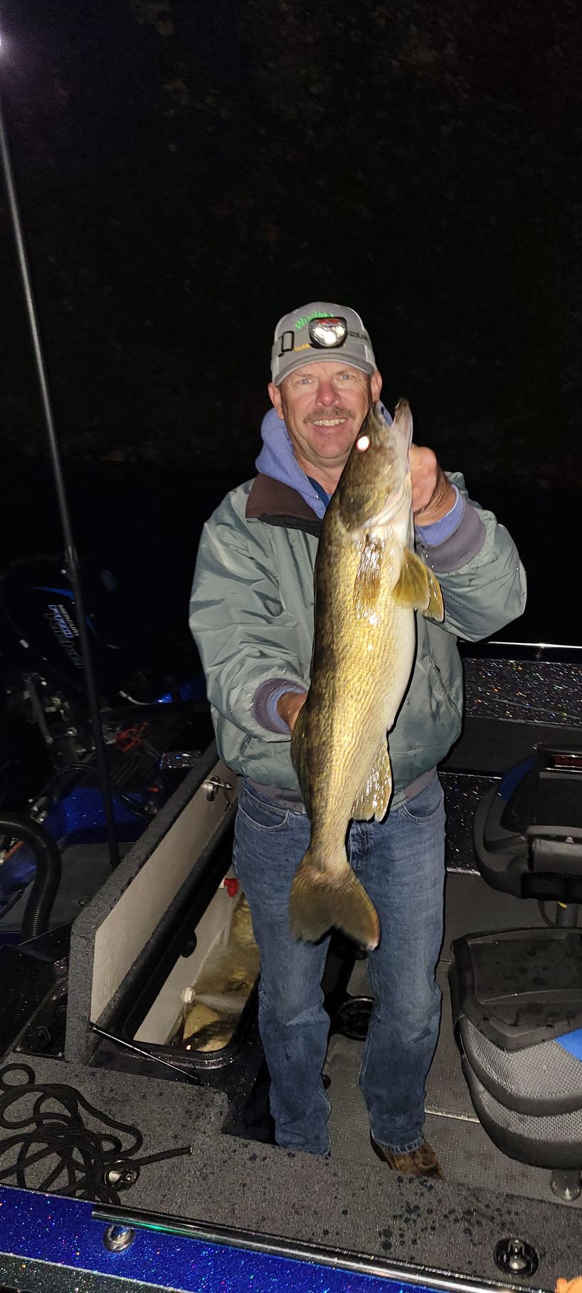 Person on a boat at night holding a large fish, wearing a gray jacket and headlamp.