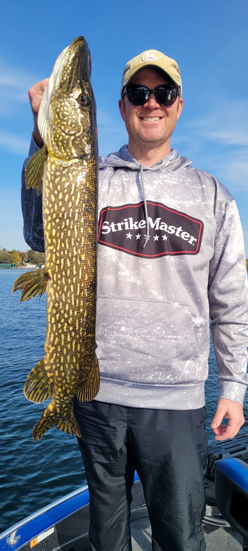 Angler holding a large northern pike on a boat, wearing sunglasses and a gray hoodie.